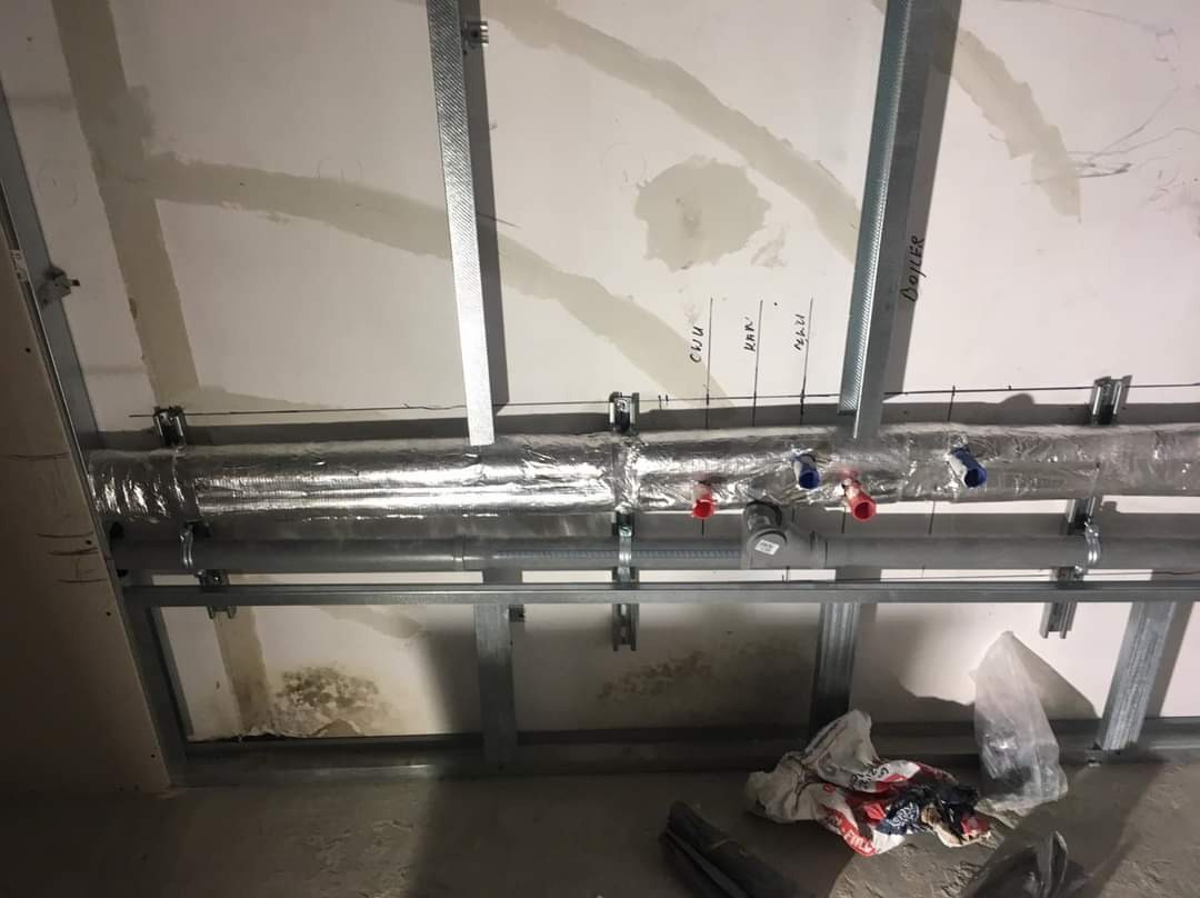 Instalacja rur grzewczych i wodnych, izolowanych termicznie, zamontowanych pod sufitem w konstrukcji z profili metalowych; widoczne oznaczenia na ścianie: CWU, HKK, NZW, BOILER.