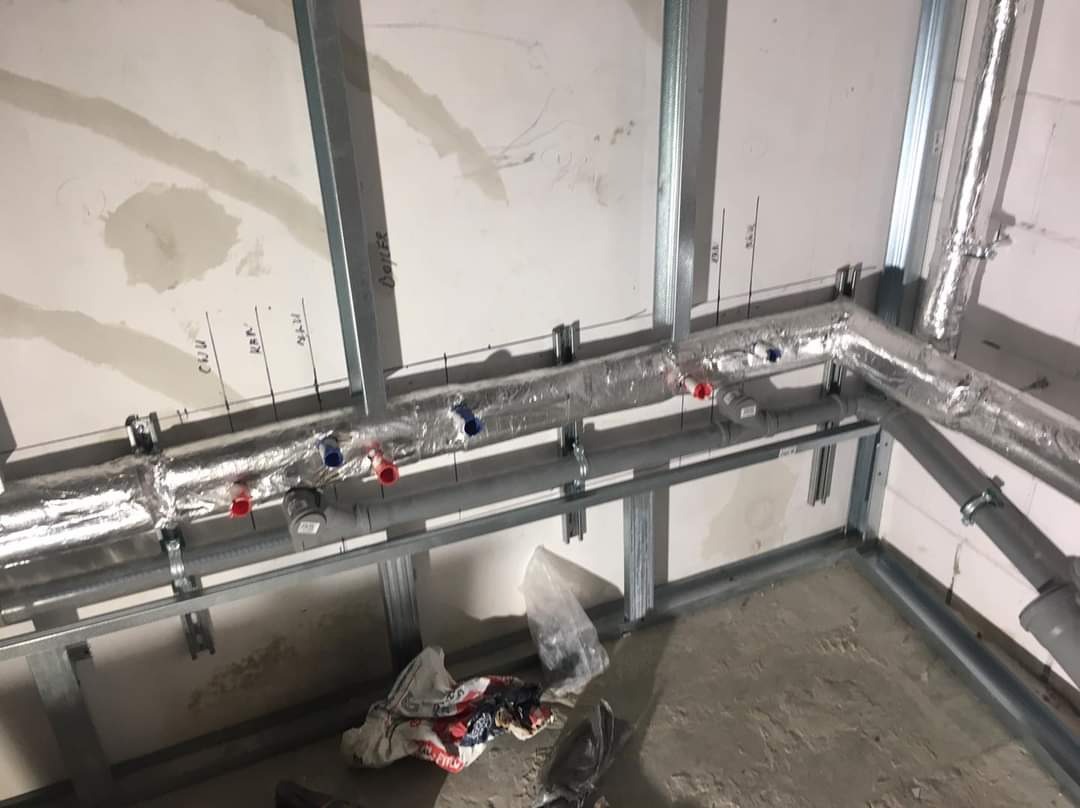 Instalacja rur grzewczych i wodnych w surowym stanie, rury izolowane folią aluminiową, widoczne oznaczenia na ścianie, konstrukcja metalowa pod zabudowę.