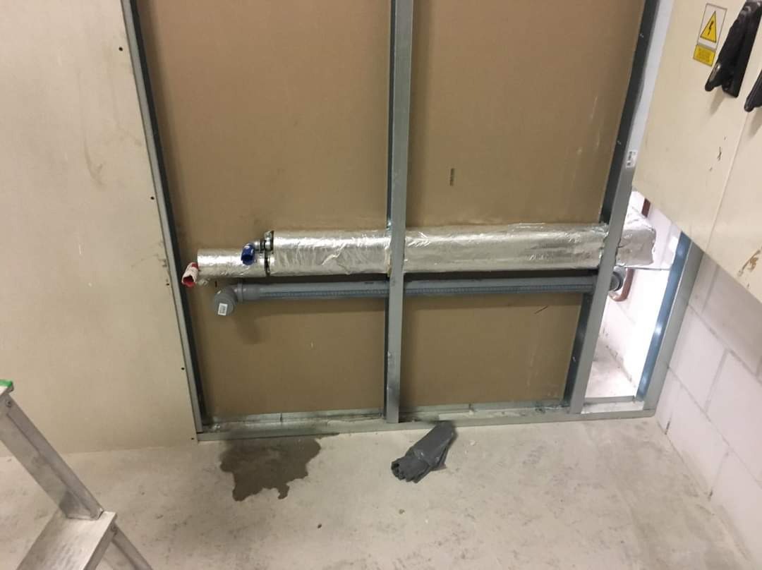 Instalacja rur w izolacji aluminiowej, widoczna wewnątrz metalowej konstrukcji ścianki działowej, z fragmentem drabiny i ciemną plamą na surowej podłodze.