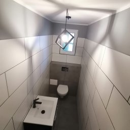 Dariusz Łokietek - Wąska toaleta wykończona białymi, prostokątnymi płytkami z ciemną fugą, szara ściana za toaletą, wisząca czarna szafka z białą umywalką i czarną armaturą, nowoczesna lampa wisząca z geometrycznym...