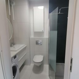 Dariusz Łokietek - Nowoczesna, jasna łazienka z pralką pod umywalką, wiszącą szafką, toaletą i kabiną prysznicową z czarną mozaiką na ścianie.