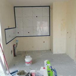 Dariusz Łokietek - Wnętrze w trakcie prac wykończeniowych: częściowo ułożone białe płytki imitujące marmur na ścianie, zabezpieczone niebieską taśmą, widoczne narzędzia i materiały budowlane, w tym wiadro z farbą...