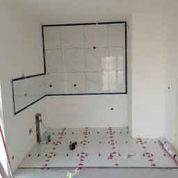 Dariusz Łokietek - Wnętrze w trakcie układania marmurowych płytek na podłodze i ścianie, z użyciem czerwonych klinów poziomujących, widoczne narzędzia i worki z zaprawą.