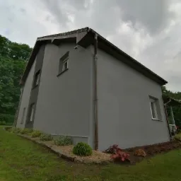 Dom jednorodzinny po tynkowaniu elewacji na szaro, z widocznymi rynnami i fragmentem ogrodu z roślinami ozdobnymi.