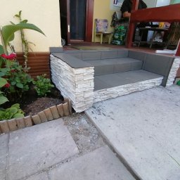 Dariusz Łokietek - Wykończone schody wejściowe do domu z szarego betonu i okładziny z jasnego kamienia dekoracyjnego, widok z poziomu gruntu, obok rabata z roślinami.