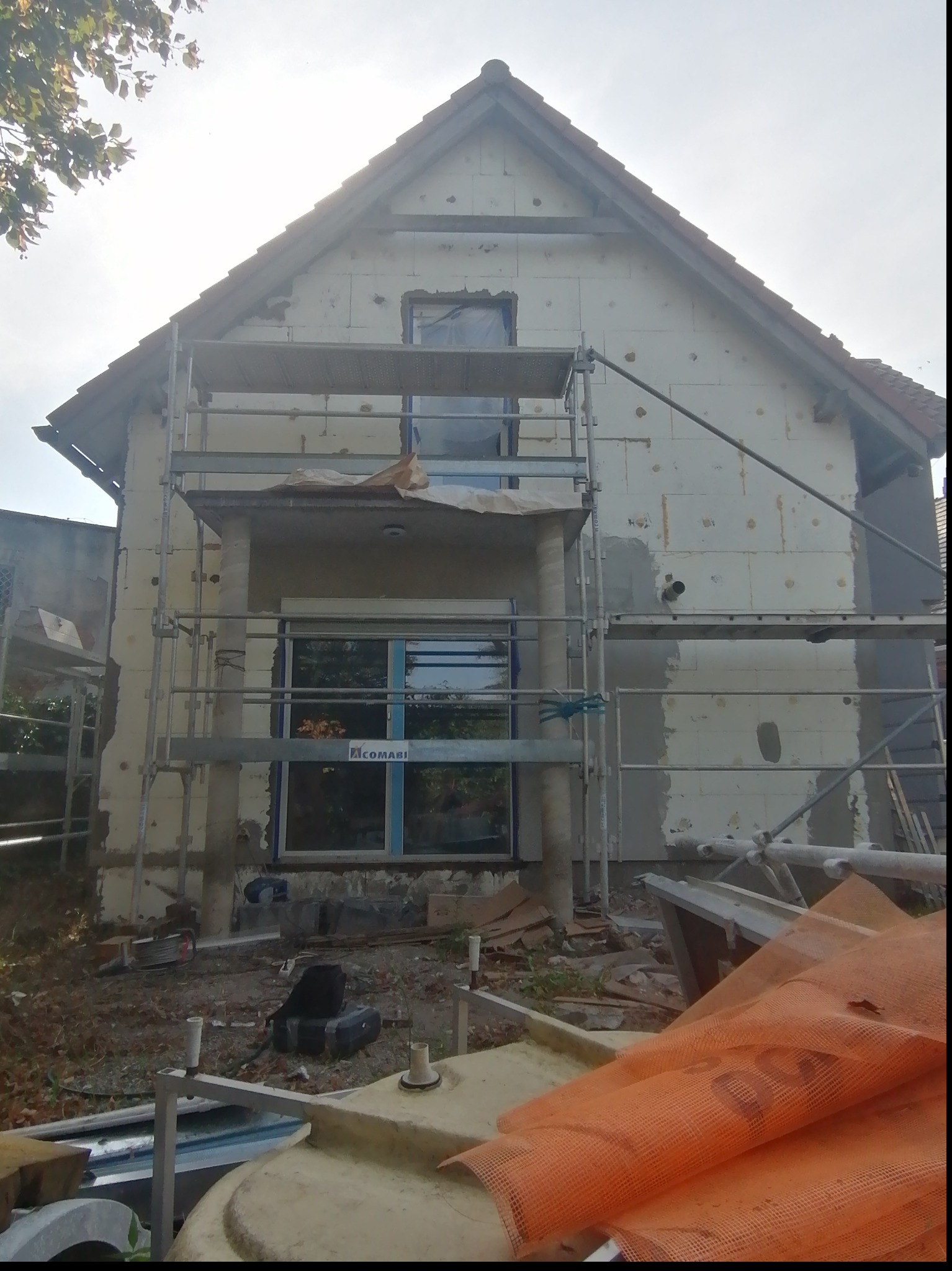 Dom jednorodzinny w trakcie ocieplania, widoczne rusztowania, styropianowe płyty izolacyjne oraz częściowo nałożona warstwa kleju z siatką, pomarańczowa siatka zabezpieczająca leży na ziemi.