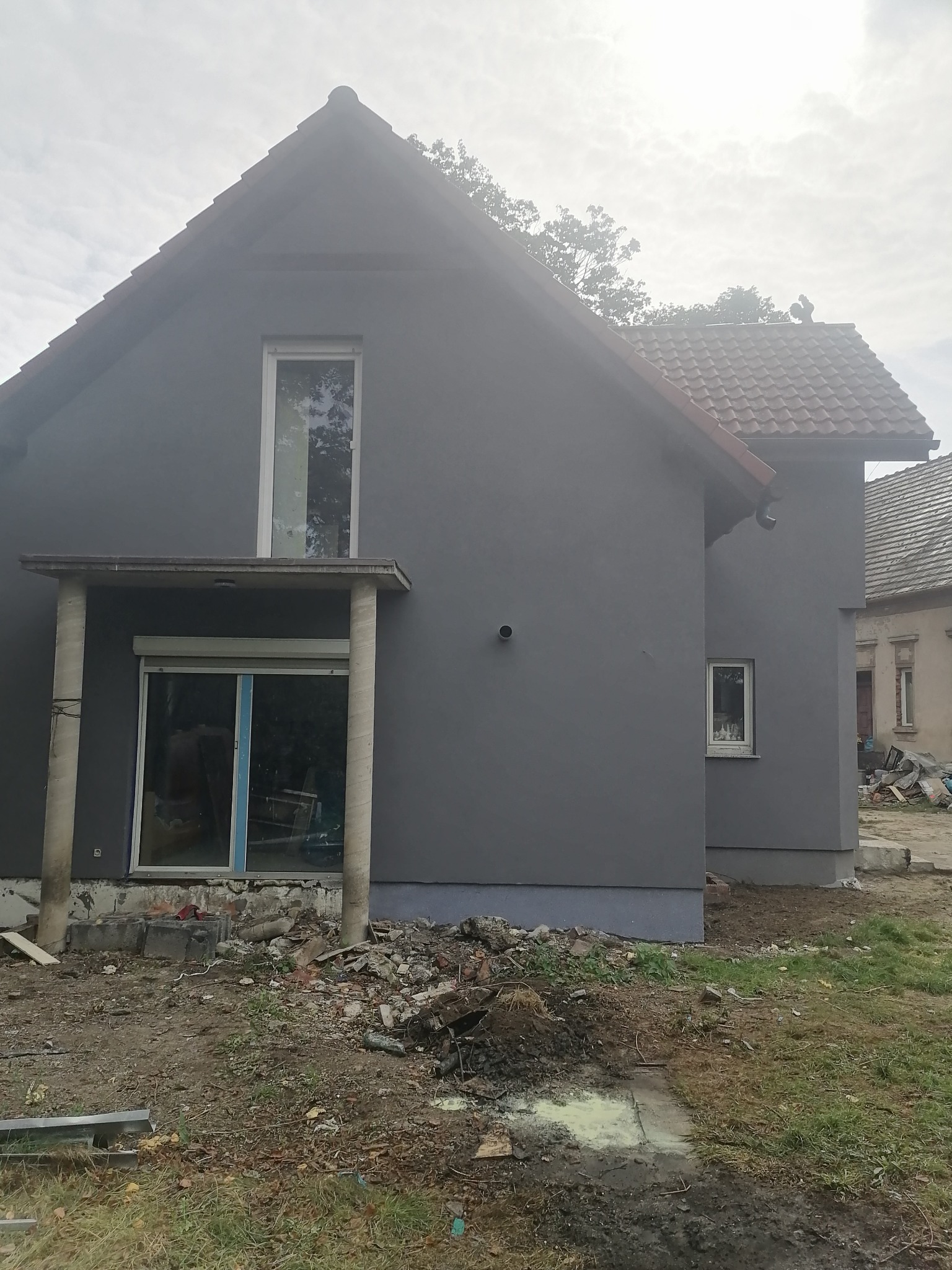 Dom jednorodzinny z szarą elewacją po tynkowaniu, widoczne ślady prac ziemnych i gruz wokół budynku, okna z częściowo zdjętą folią ochronną.