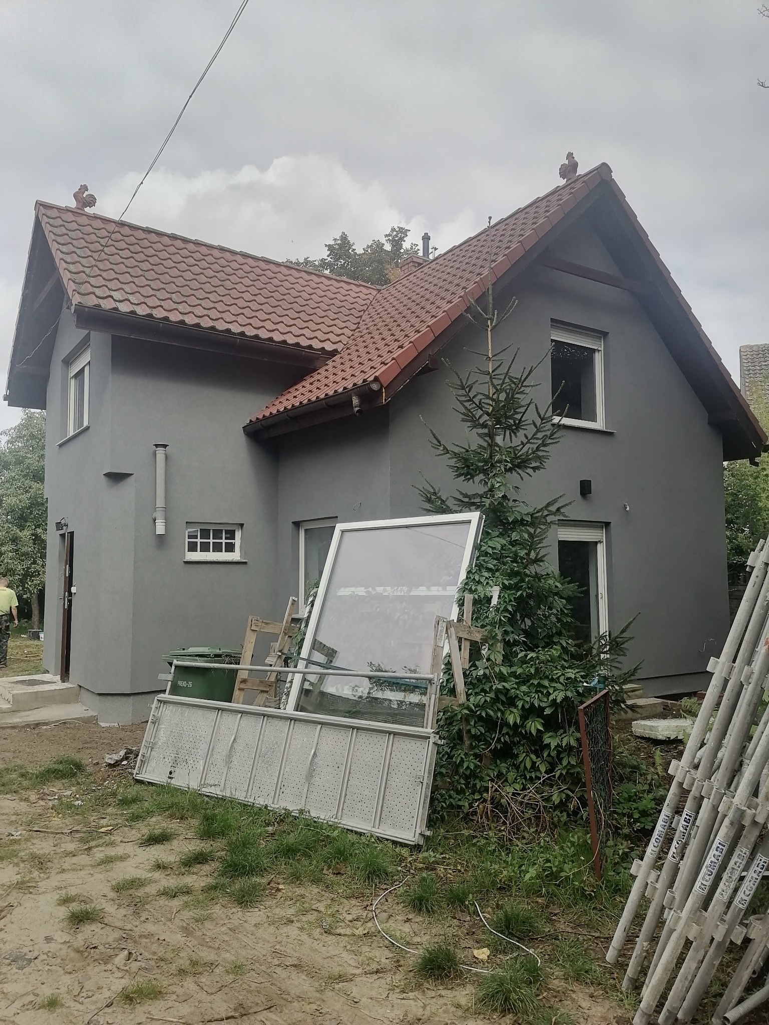 Dwupiętrowy dom z szarą elewacją i czerwoną dachówką, z widocznymi rusztowaniami i elementami po remoncie, takimi jak okno i drabina, a na dachu ozdobne koguty.