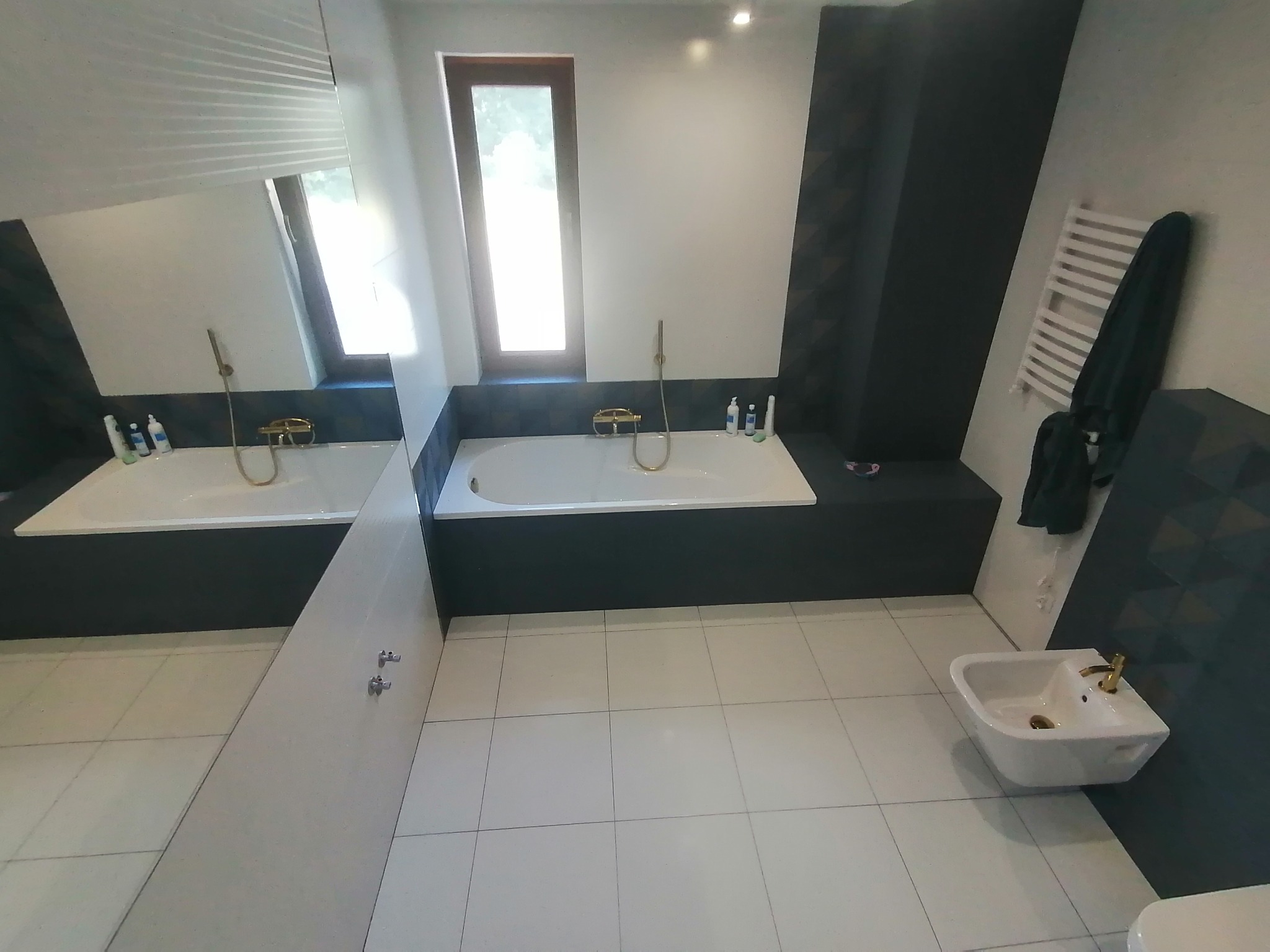 Nowoczesna łazienka z białą wanną, bidetem z złotą armaturą, grzejnikiem drabinkowym i ciemnymi panelami dekoracyjnymi na ścianach.