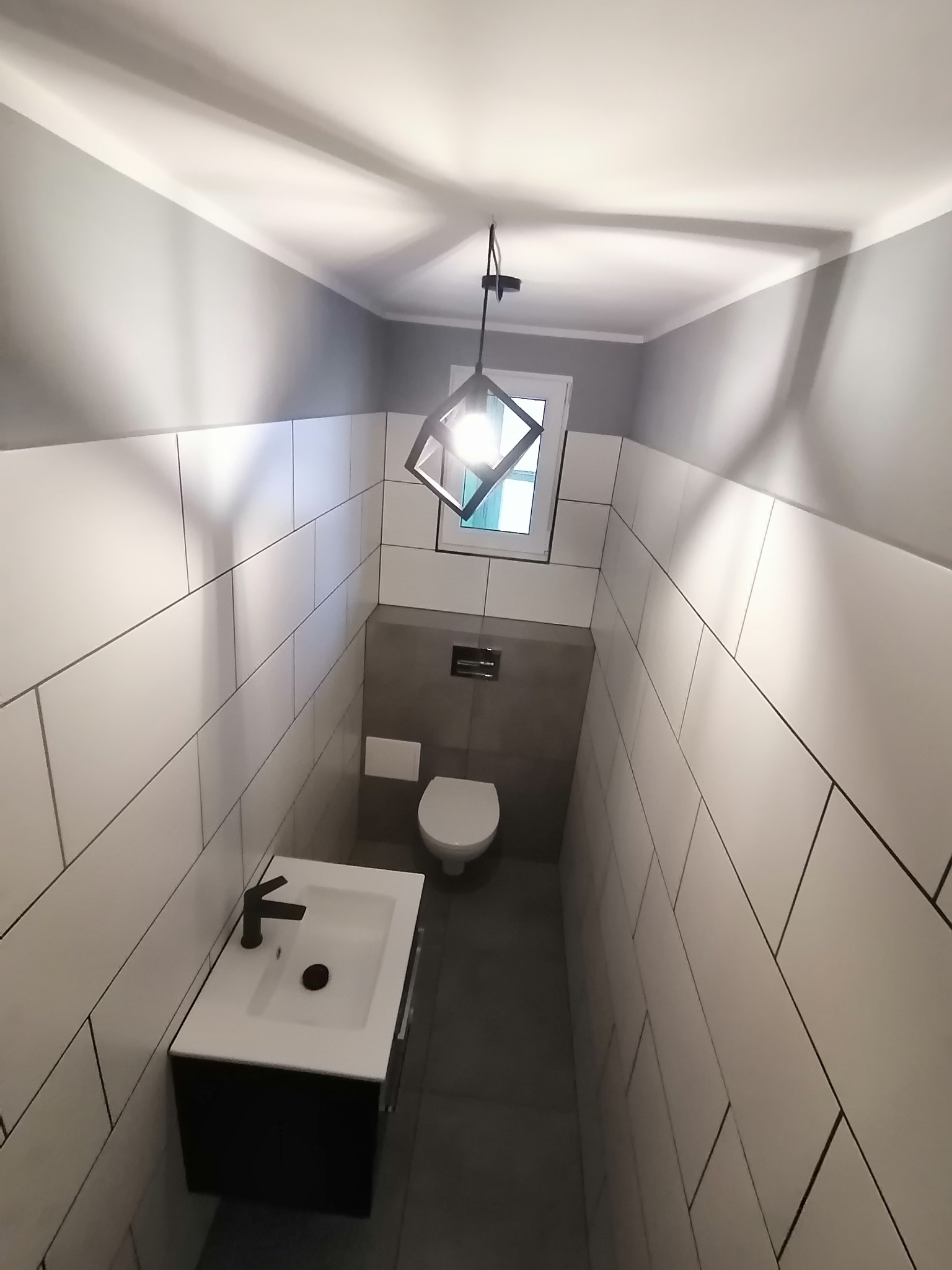 Wąska toaleta wykończona białymi, prostokątnymi płytkami z ciemną fugą, szara ściana za toaletą, wisząca czarna szafka z białą umywalką i czarną armaturą, nowoczesna lampa wisząca z geometrycznym...