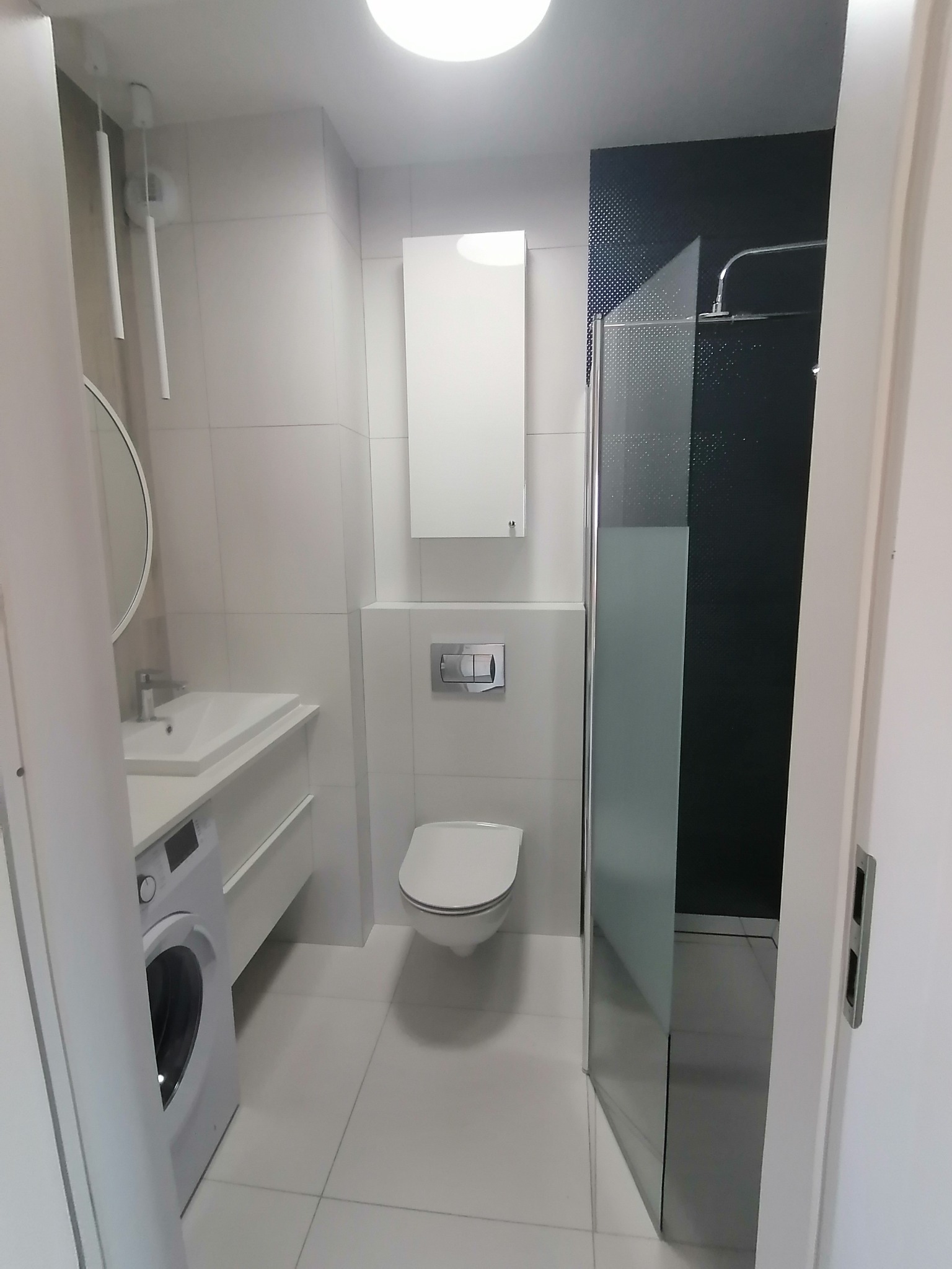 Nowoczesna, jasna łazienka z pralką pod umywalką, wiszącą szafką, toaletą i kabiną prysznicową z czarną mozaiką na ścianie.