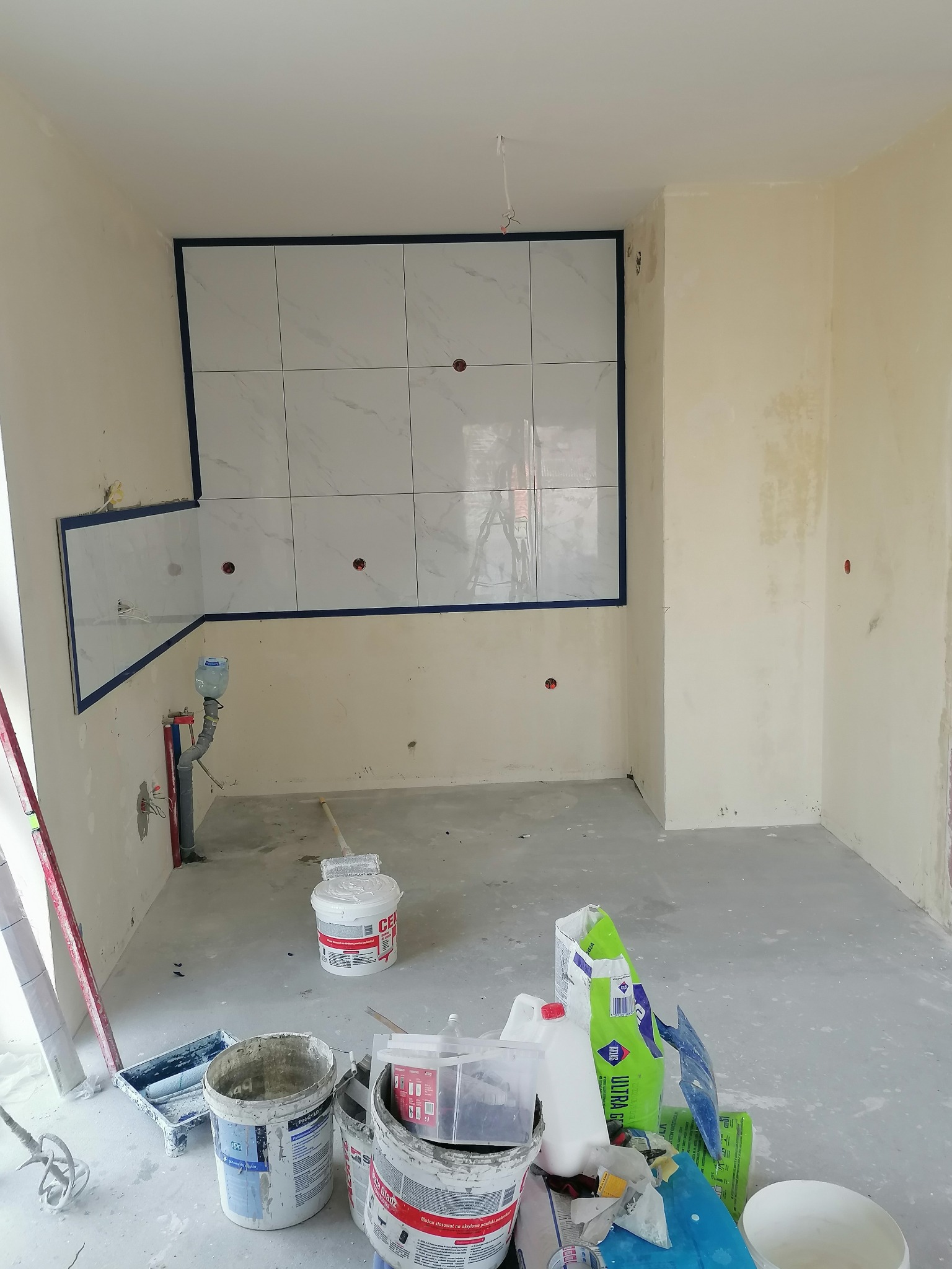 Wnętrze w trakcie prac wykończeniowych: częściowo ułożone białe płytki imitujące marmur na ścianie, zabezpieczone niebieską taśmą, widoczne narzędzia i materiały budowlane, w tym wiadro z farbą...