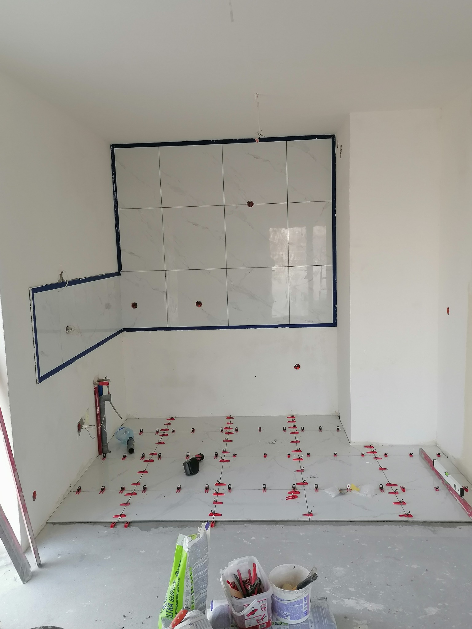 Wnętrze w trakcie układania marmurowych płytek na podłodze i ścianie, z użyciem czerwonych klinów poziomujących, widoczne narzędzia i worki z zaprawą.