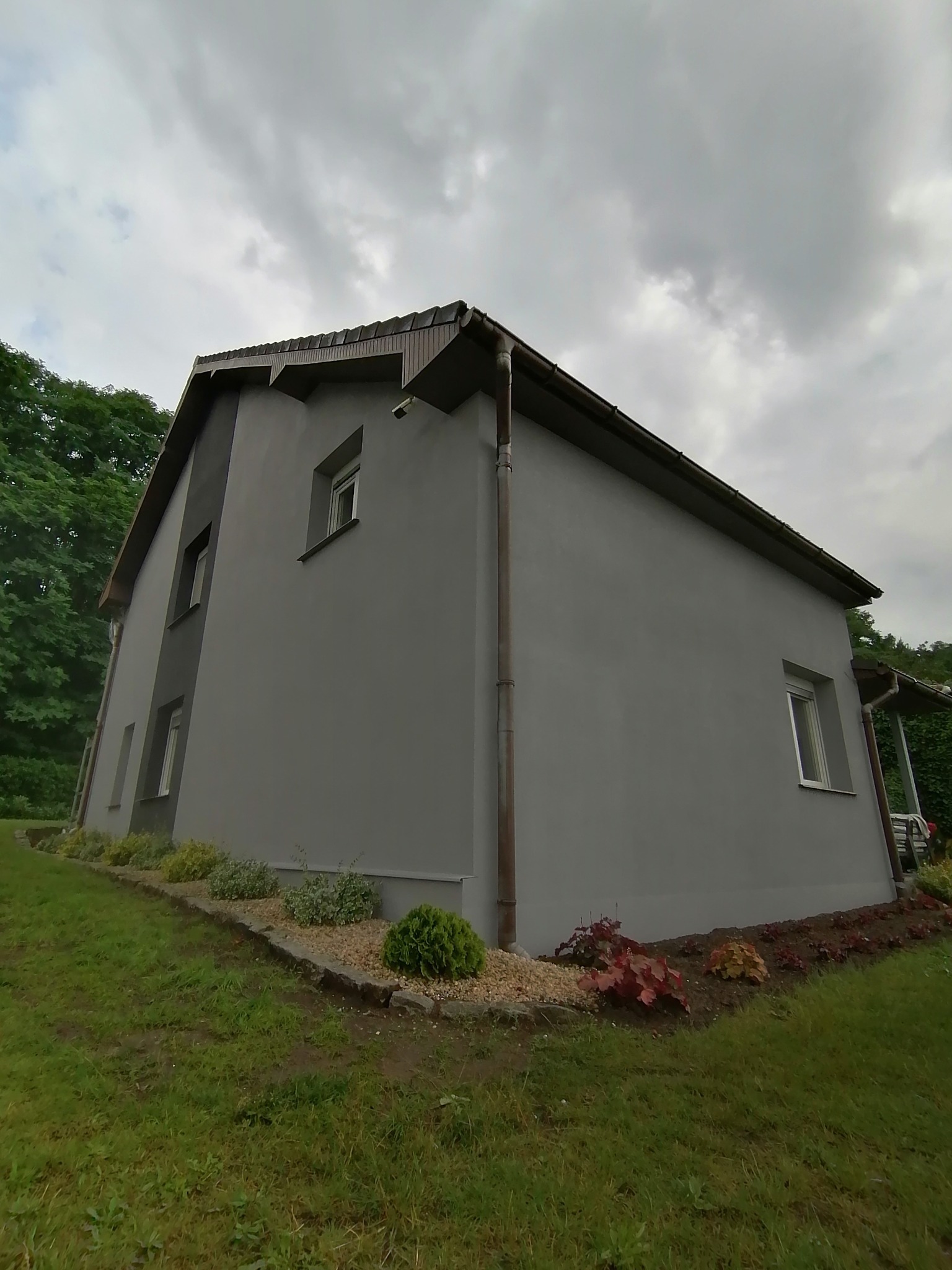 Dom jednorodzinny po tynkowaniu elewacji na szaro, z widocznymi rynnami i fragmentem ogrodu z roślinami ozdobnymi.