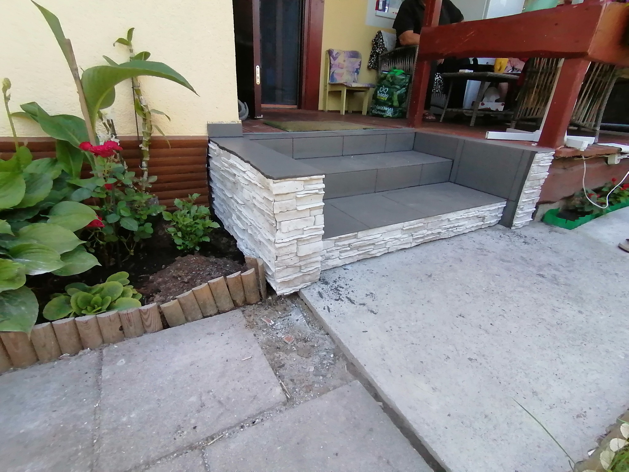 Wykończone schody wejściowe do domu z szarego betonu i okładziny z jasnego kamienia dekoracyjnego, widok z poziomu gruntu, obok rabata z roślinami.