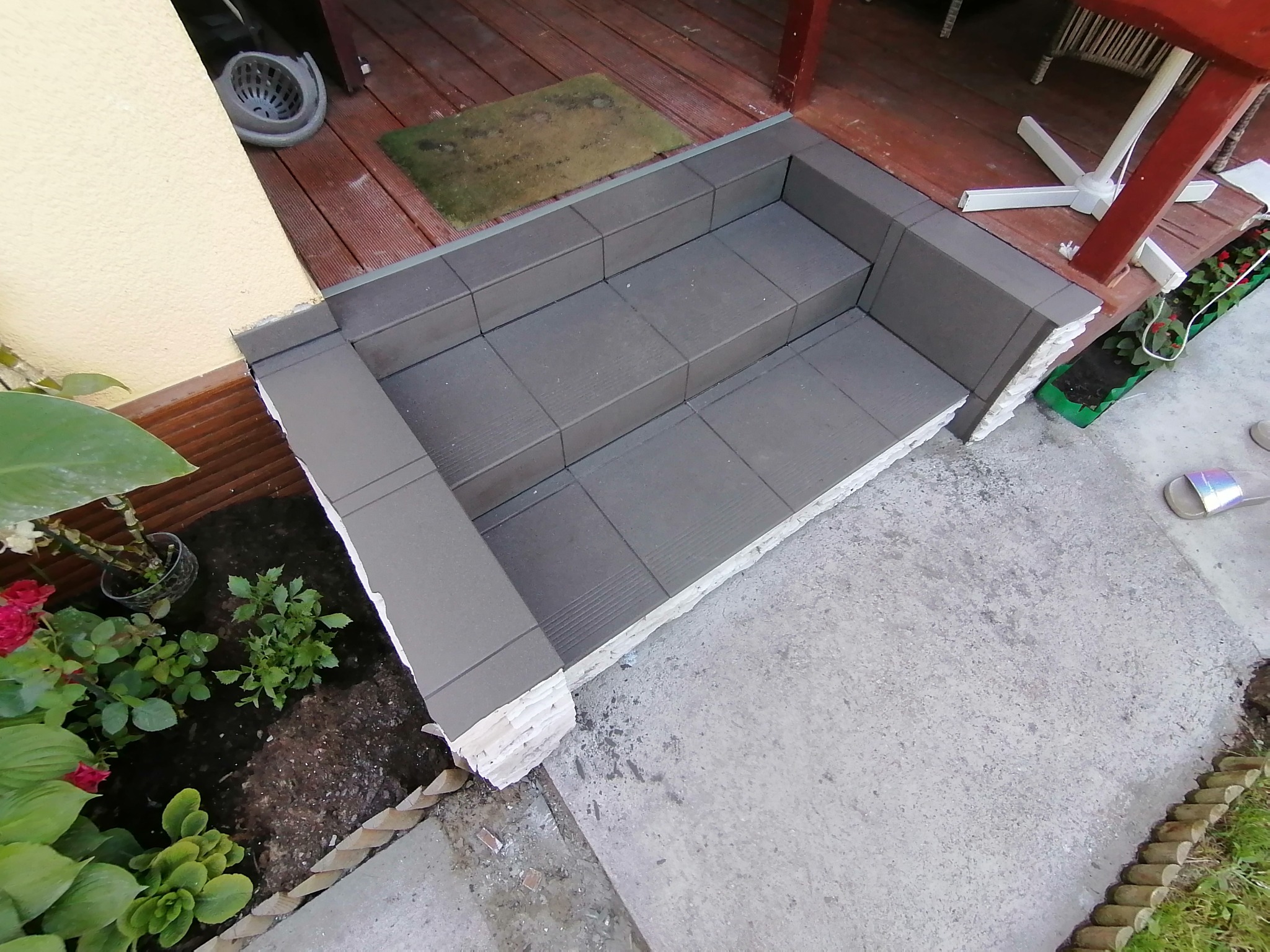 Wykończone schody wejściowe z szarych płytek, z widocznym ociepleniem styropianowym i fragmentem drewnianego tarasu oraz roślinami w donicach po bokach.