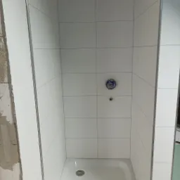 Biała kabina prysznicowa z odpływem w posadzce, wyłożona kwadratowymi płytkami, widoczna w trakcie remontu łazienki w Koszalinie, z zabezpieczonym żółtą taśmą górnym rogiem.