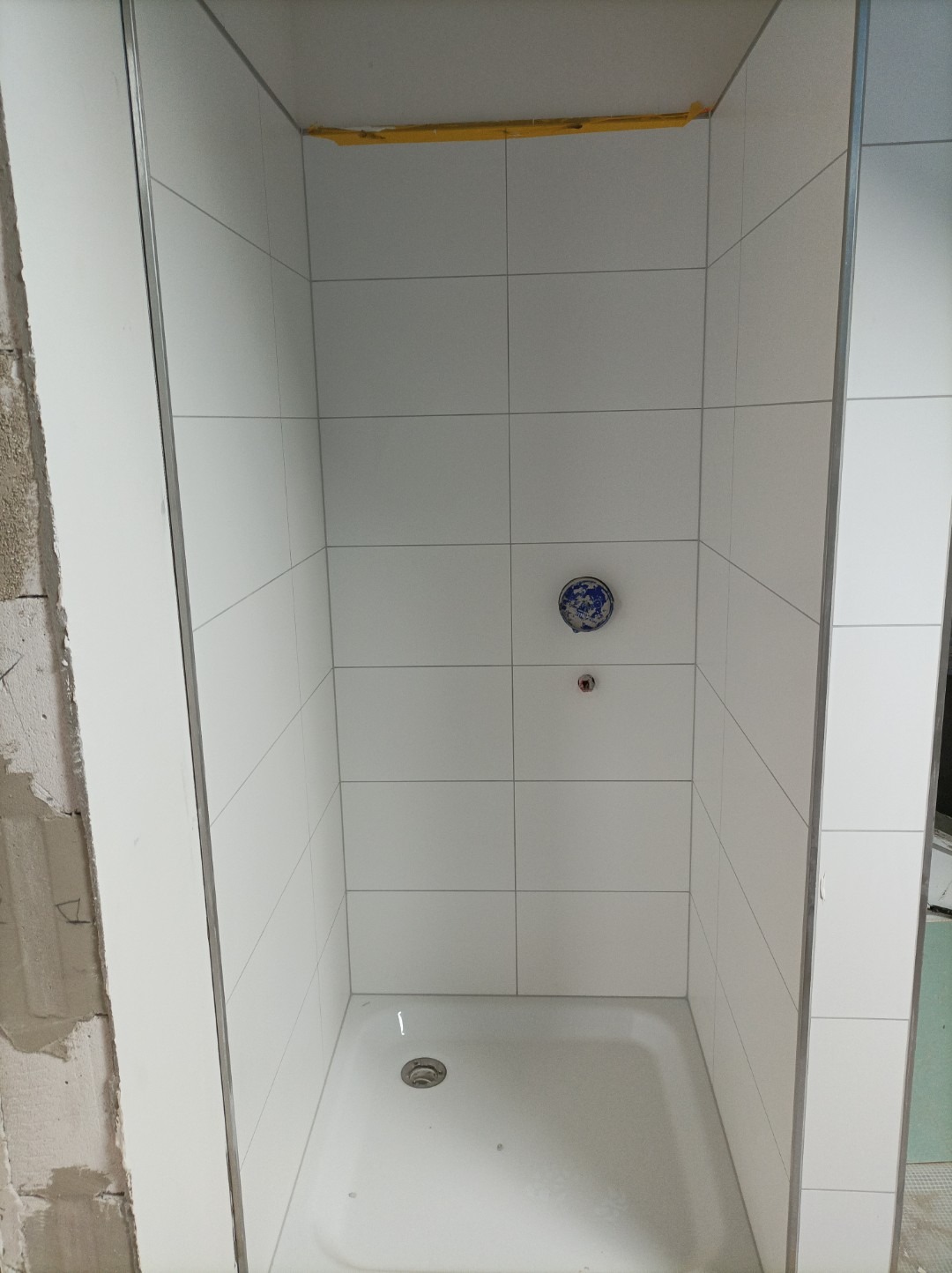 Biała kabina prysznicowa z odpływem w posadzce, wyłożona kwadratowymi płytkami, widoczna w trakcie remontu łazienki w Koszalinie, z zabezpieczonym żółtą taśmą górnym rogiem.