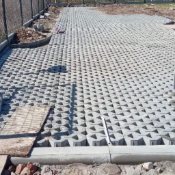 Szara, ażurowa kostka betonowa ułożona na przygotowanym podłożu, widoczne obrzeża betonowe i fragment ogrodzenia panelowego z bramką, na części powierzchni kostka jeszcze nieułożona.