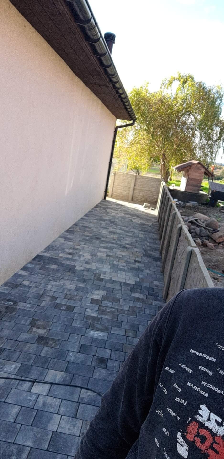 Ukończona ścieżka z szarej kostki brukowej, biegnąca wzdłuż ściany domu i betonowego ogrodzenia, z widocznym fragmentem osoby w ubraniu.