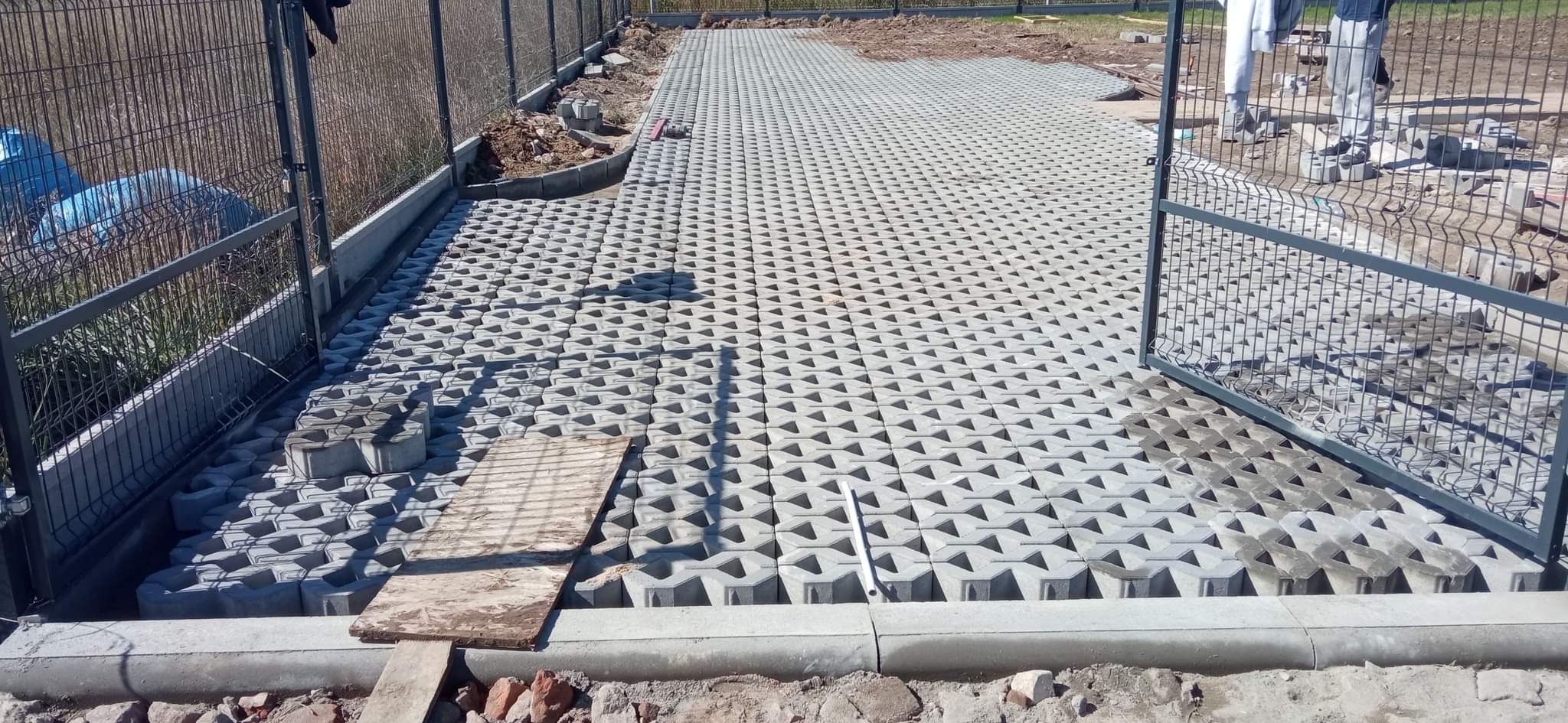 Szara, ażurowa kostka betonowa ułożona na przygotowanym podłożu, widoczne obrzeża betonowe i fragment ogrodzenia panelowego z bramką, na części powierzchni kostka jeszcze nieułożona.