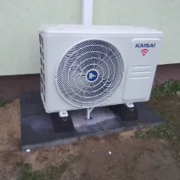 Zewnętrzna jednostka klimatyzacji Kaisai zamontowana na betonowej podstawie z czarnymi podkładkami antywibracyjnymi, widoczne przyłącze rurowe, trawa i elewacja budynku w tle.