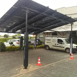 Zadaszenie parkingu z paneli fotowoltaicznych, obok biały samochód dostawczy z logo Digiset, pomarańczowy pachołek, stanowiska do ładowania aut elektrycznych.