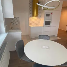 Nowoczesna, minimalistyczna kuchnia z białymi szafkami, złotą armaturą i okrągłym stołem z szarymi krzesłami. Na blacie leżą zapakowane próbki. Charakterystyczny złoty okap i designerska lampa...