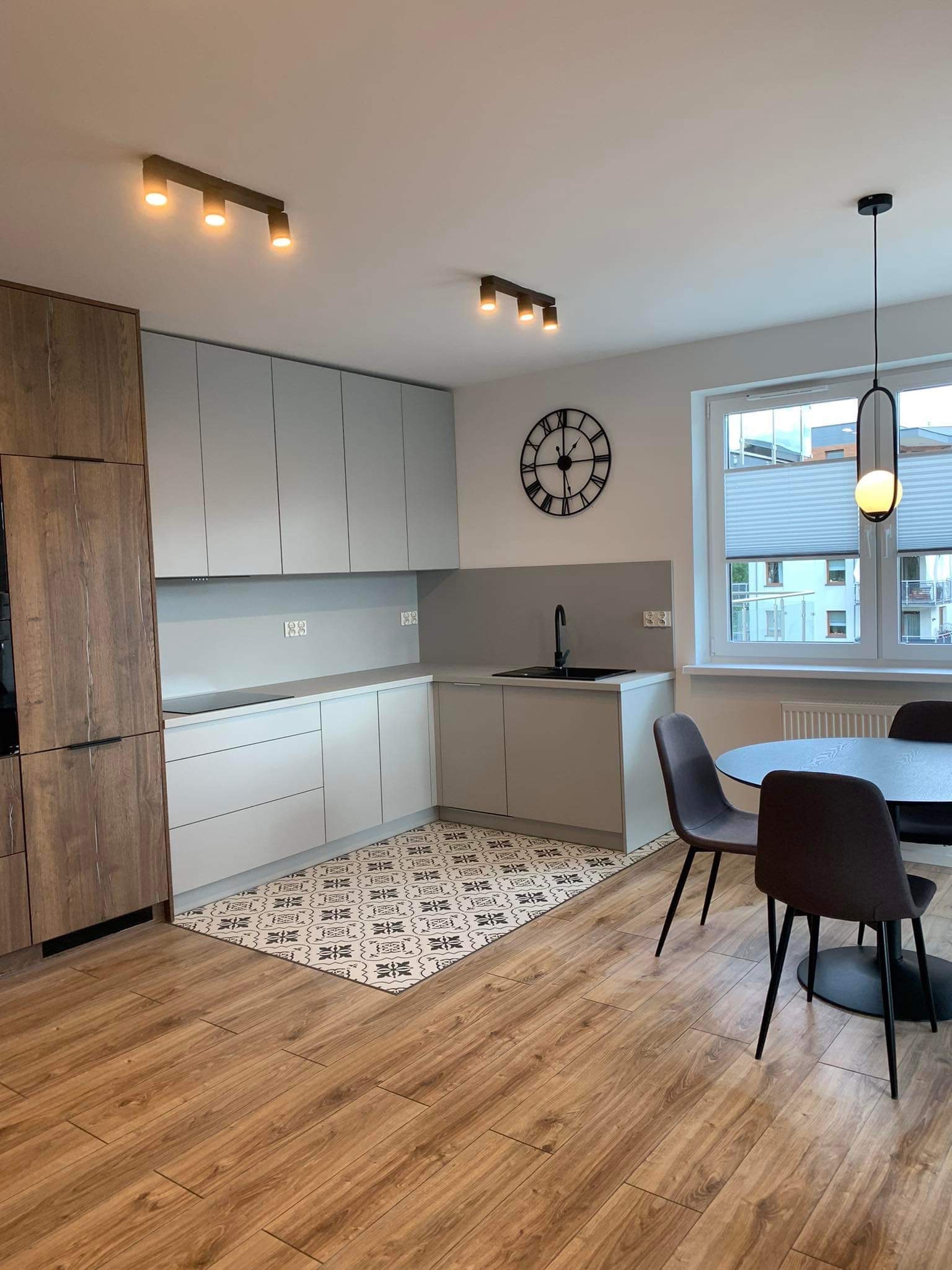 Nowoczesna kuchnia z szarymi szafkami, drewnianymi akcentami i wzorzystą podłogą, oświetlona lampami sufitowymi i wiszącą lampą nad okrągłym stołem z krzesłami.