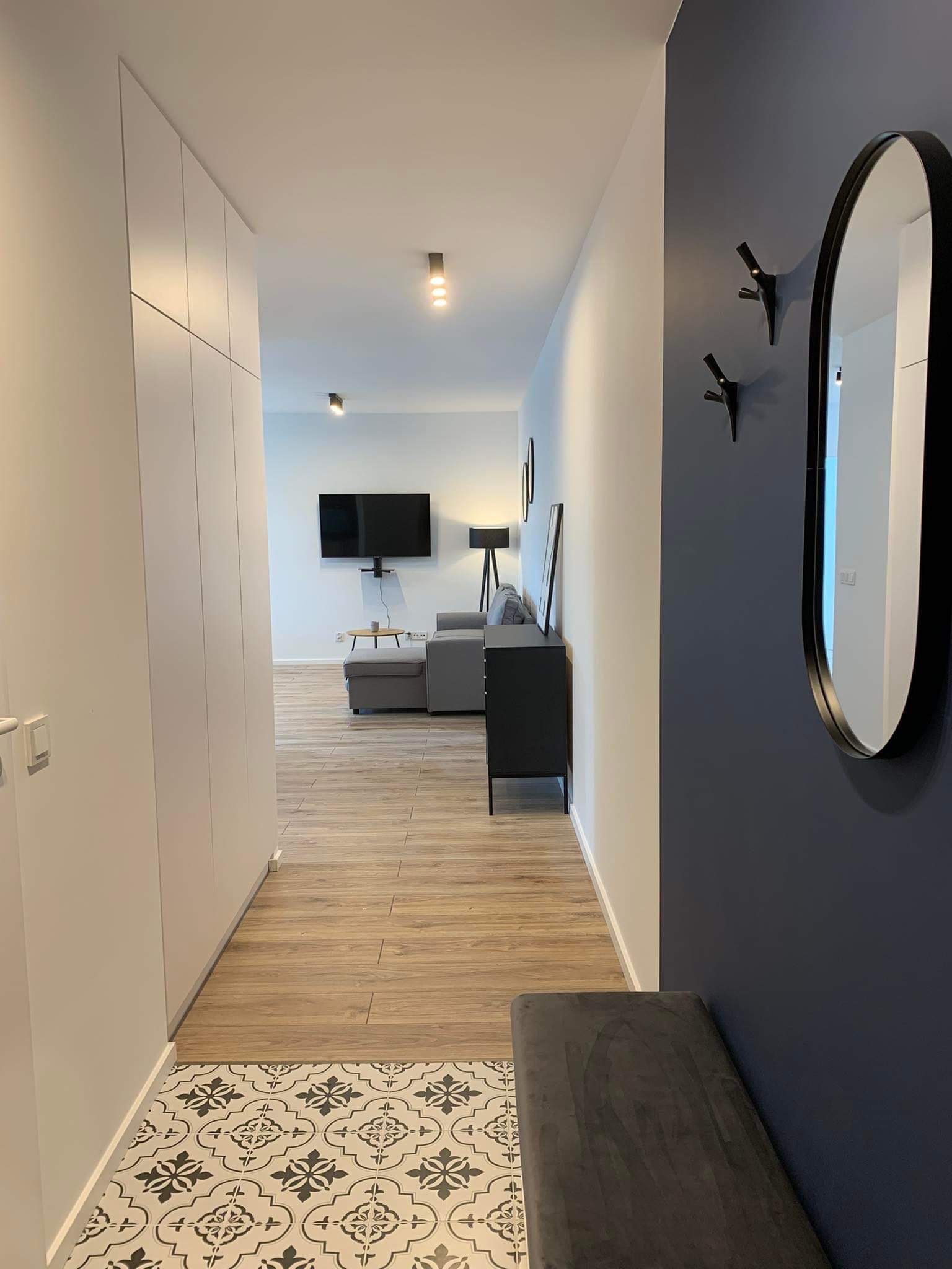 Nowoczesny przedpokój z białą szafą w zabudowie, drewnopodobną podłogą, ozdobnymi płytkami, ciemną ścianą z wieszakami w kształcie gałęzi i owalnym lustrem.