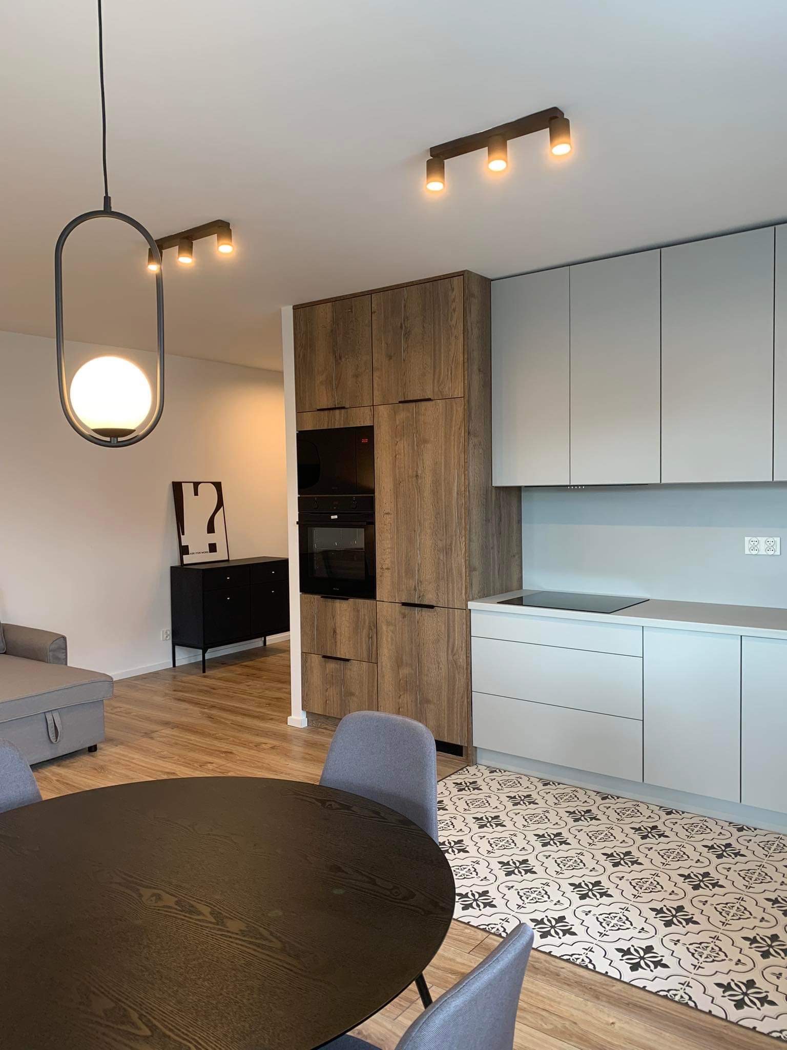 Nowoczesna kuchnia z drewnianą zabudową piekarnika i szarymi szafkami, okrągły stół z krzesłami i wzorzysta podłoga w otwartej przestrzeni.