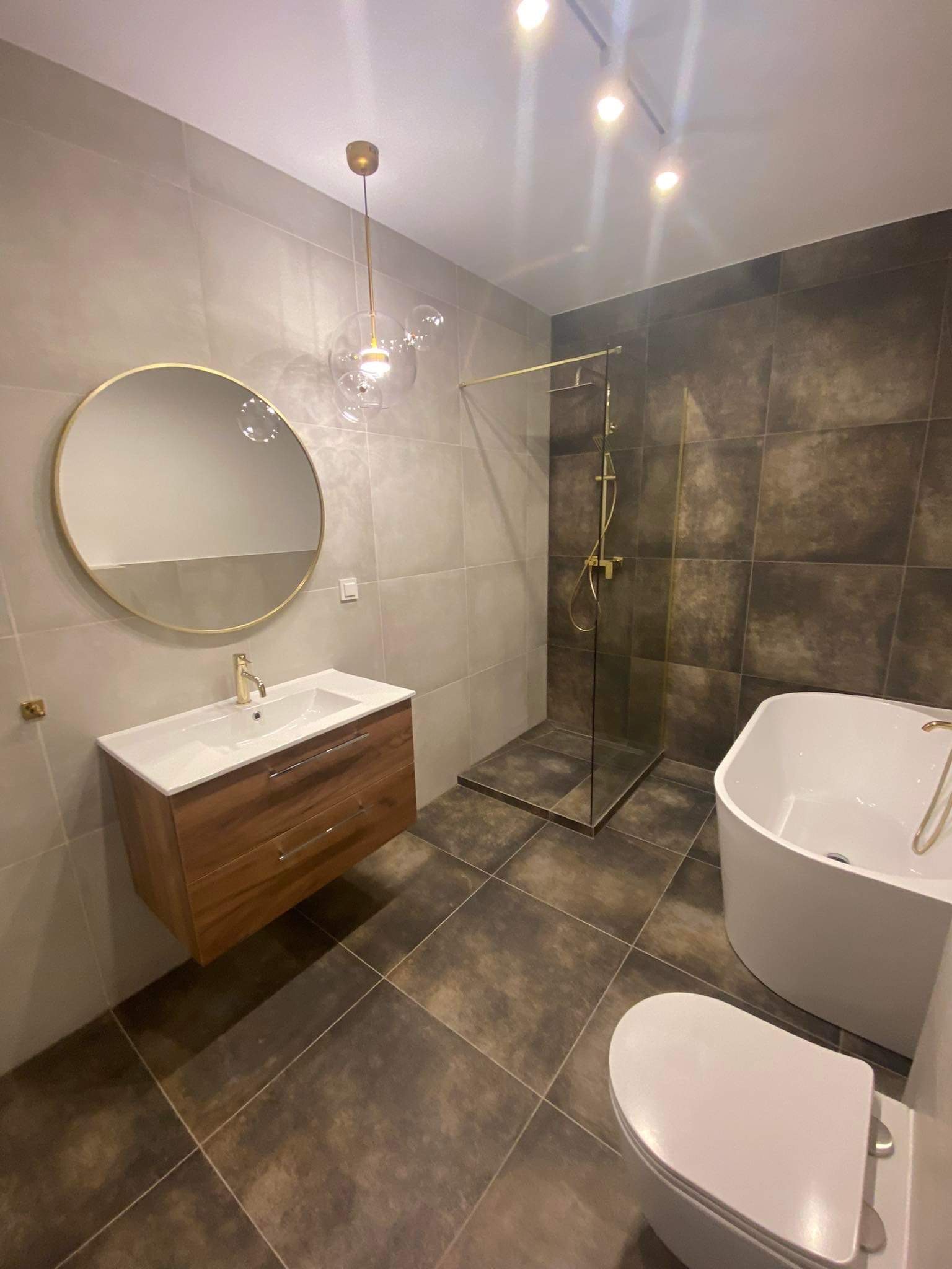 Nowoczesna łazienka z brązowymi płytkami, kabiną prysznicową ze złotymi elementami, okrągłym lustrem i drewnianą szafką pod umywalką oraz designerską lampą wiszącą.