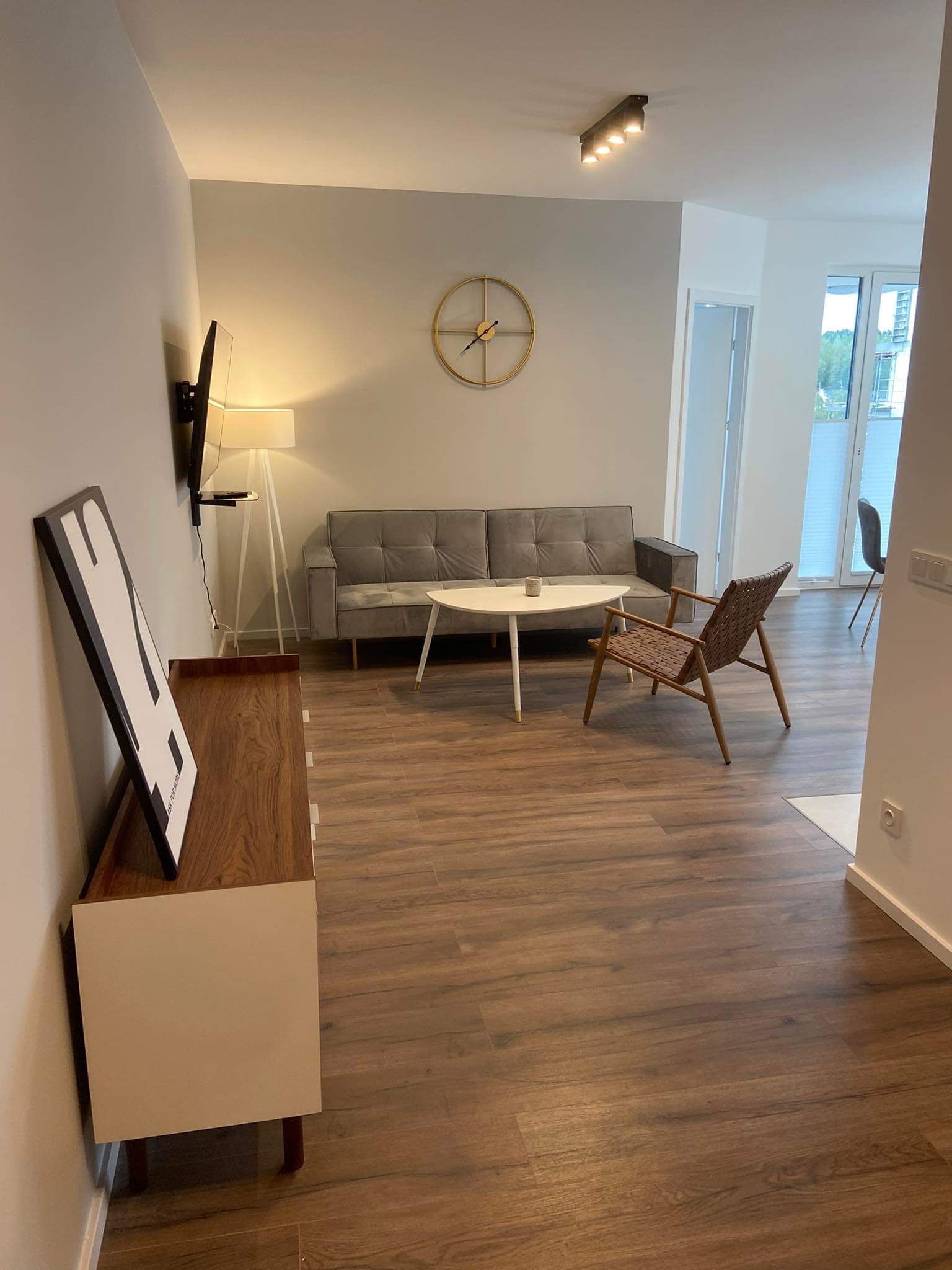 Jasne wnętrze pokoju dziennego z drewnianą podłogą, szarą sofą, białym stolikiem kawowym, designerskim krzesłem z plecionki, szafką RTV z plakatem i złotym zegarem na ścianie.