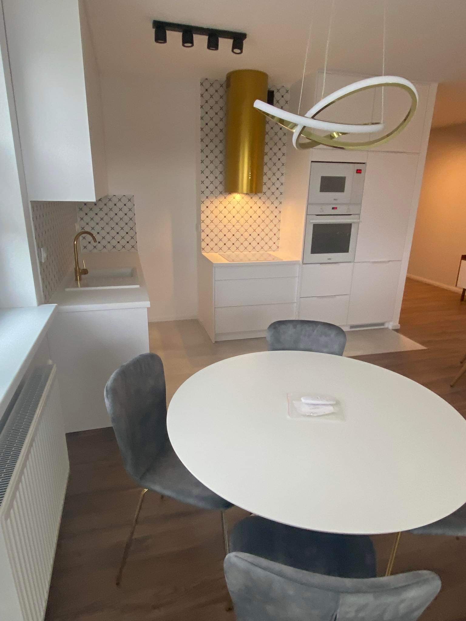 Nowoczesna, minimalistyczna kuchnia z białymi szafkami, złotą armaturą i okrągłym stołem z szarymi krzesłami. Na blacie leżą zapakowane próbki. Charakterystyczny złoty okap i designerska lampa...
