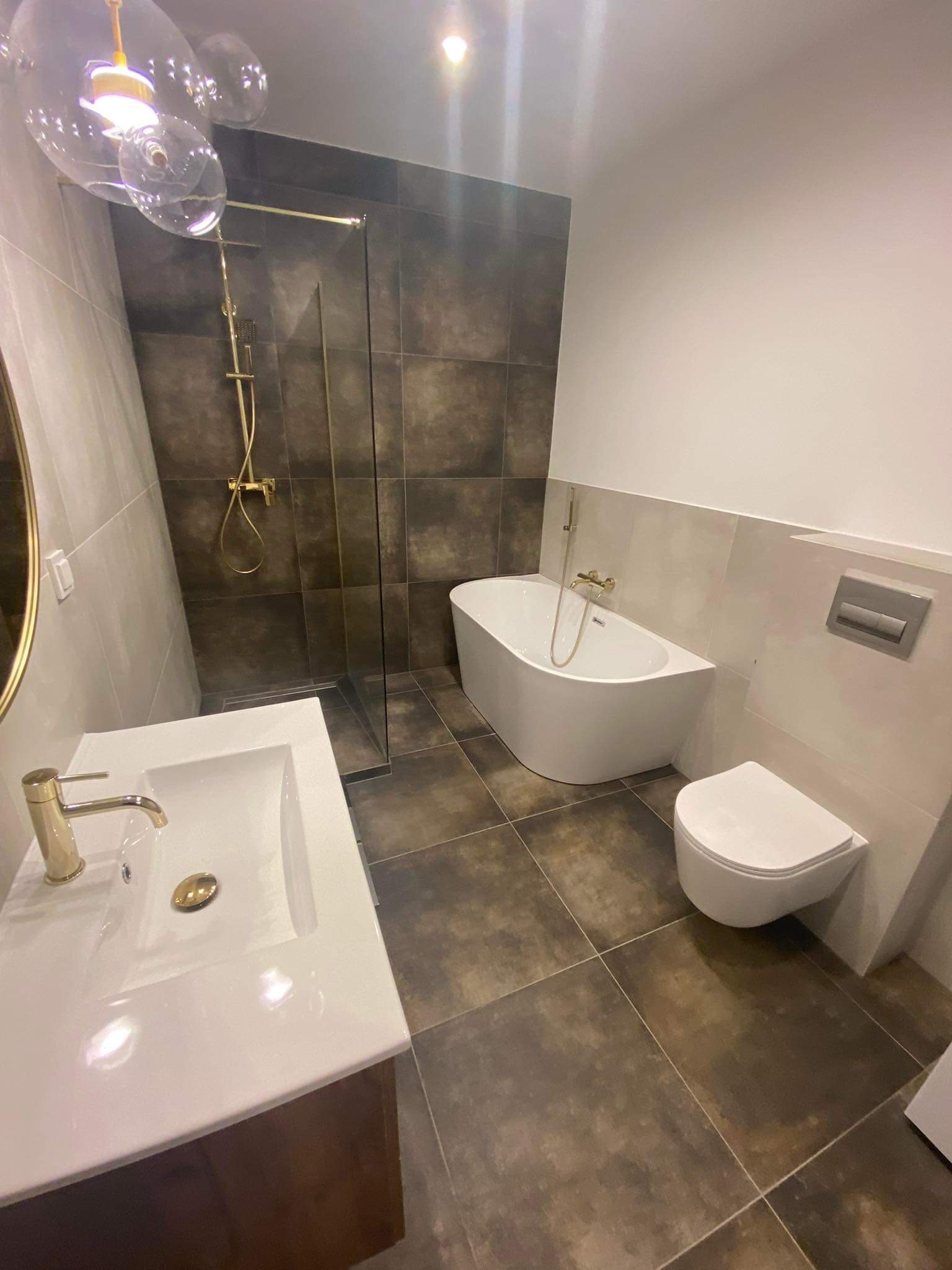 Nowoczesna łazienka z brązowymi płytkami, kabiną prysznicową typu walk-in ze złotą armaturą, białą wanną wolnostojącą i umywalką nablatową na szafce z ciemnego drewna, oświetlona dekoracyjną lampą...