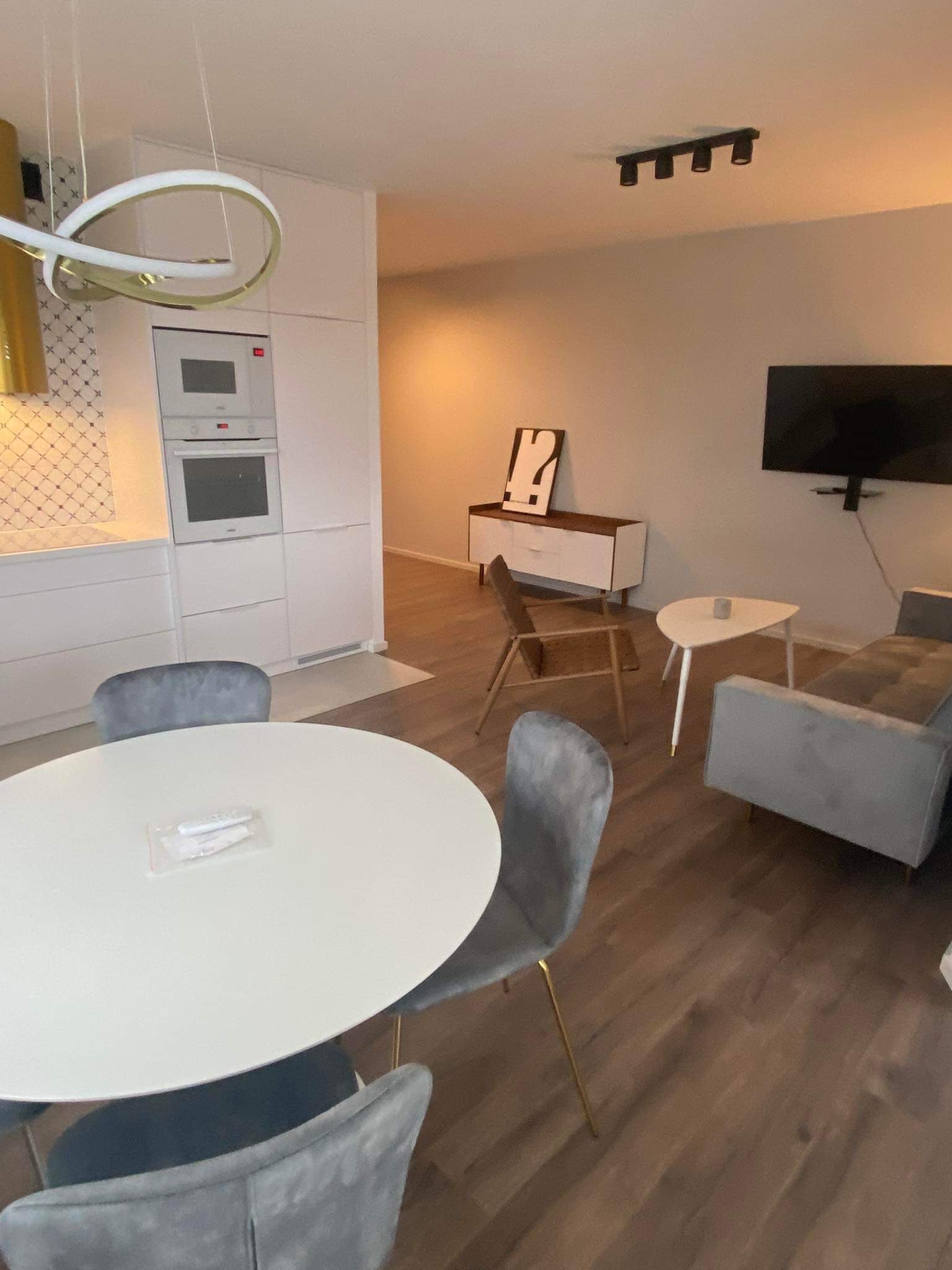 Jasne wnętrze mieszkania z aneksem kuchennym, okrągłym stołem z szarymi krzesłami, szarą sofą i drewnianą podłogą. Na stole leżą zapakowane sztućce. W tle białe szafki kuchenne z piekarnikiem...