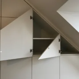 Biała szafa wnękowa pod skosem dachu z otwartymi drzwiczkami, widoczne półki wewnątrz.