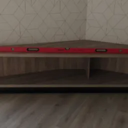 Narożna szafka RTV z drewnopodobnym wykończeniem, ustawiona na czarnej, metalowej ramie, z czerwoną poziomicą na górze, na tle ściany z geometrycznym wzorem.