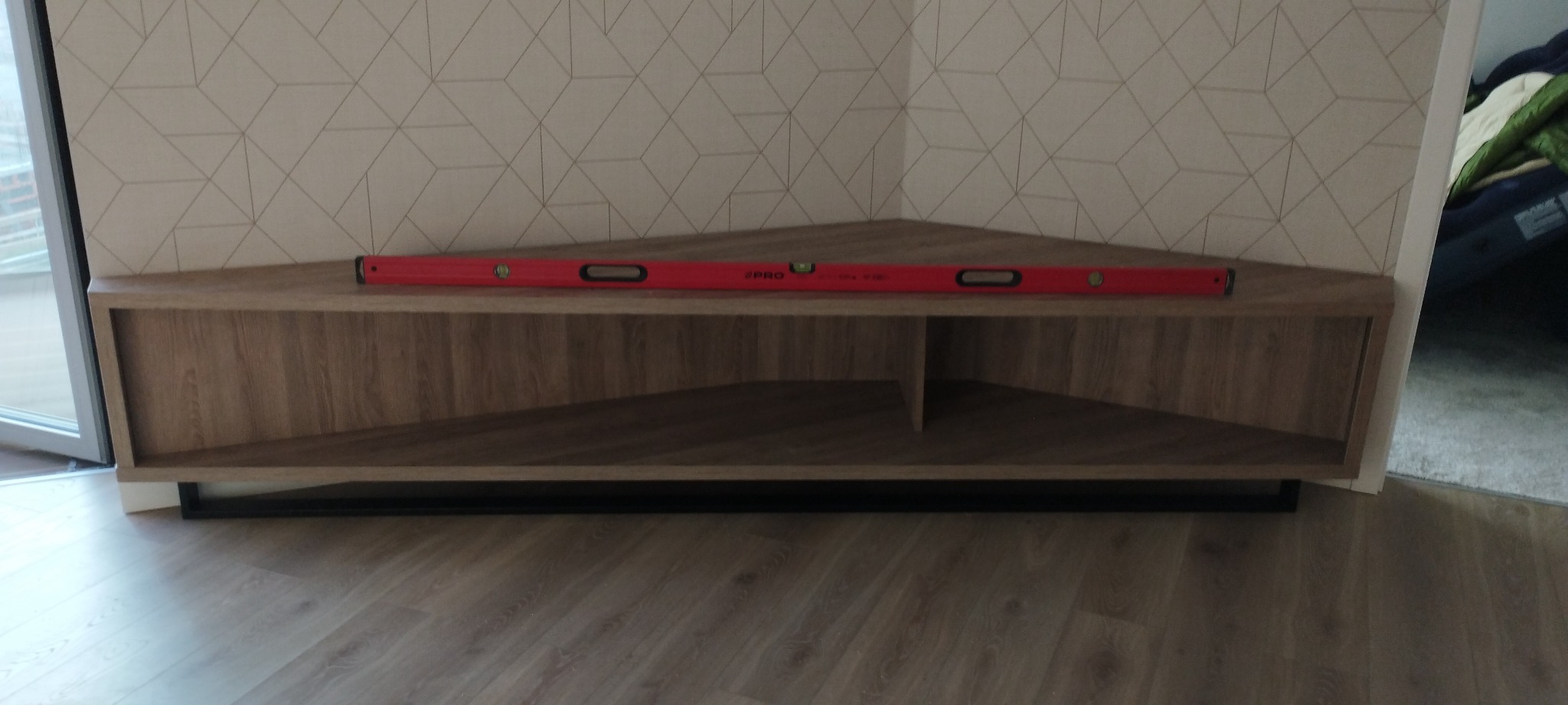 Narożna szafka RTV z drewnopodobnym wykończeniem, ustawiona na czarnej, metalowej ramie, z czerwoną poziomicą na górze, na tle ściany z geometrycznym wzorem.