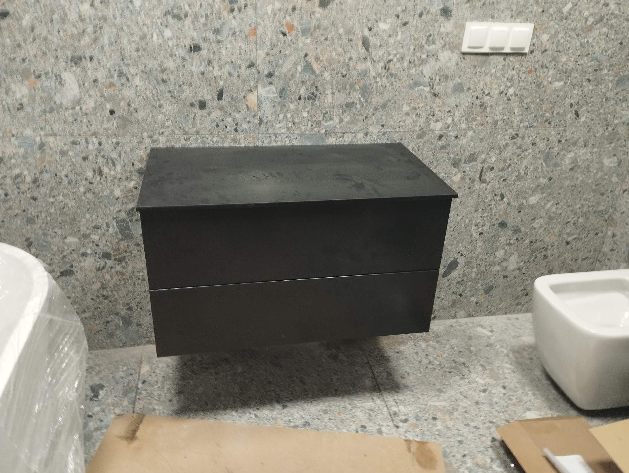 Czarna, wisząca szafka łazienkowa z dwiema szufladami, zamontowana na ścianie wyłożonej płytkami imitującymi lastryko. Obok widoczna owinięta folią wanna oraz umywalka. Na podłodze kartony po montażu.