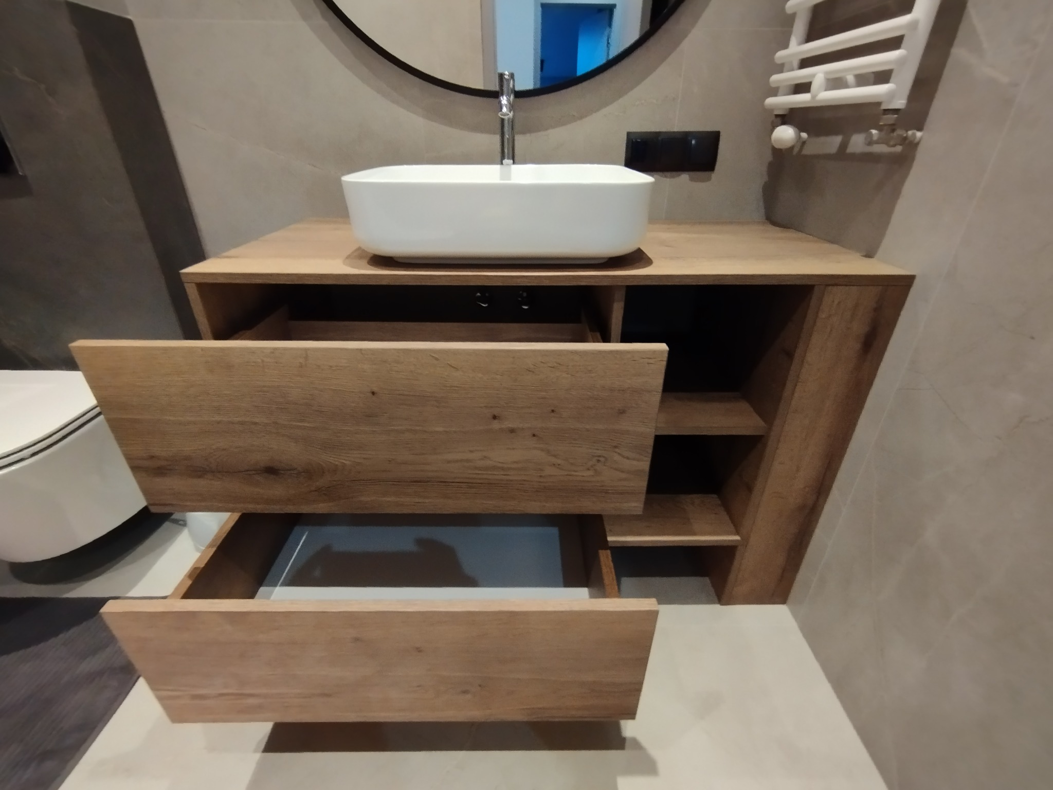 Łazienka z drewnianą szafką pod umywalką z dwoma wysuwanymi szufladami, umywalka nablatowa, okrągłe lustro, grzejnik drabinkowy i szaro-beżowe płytki na ścianach.