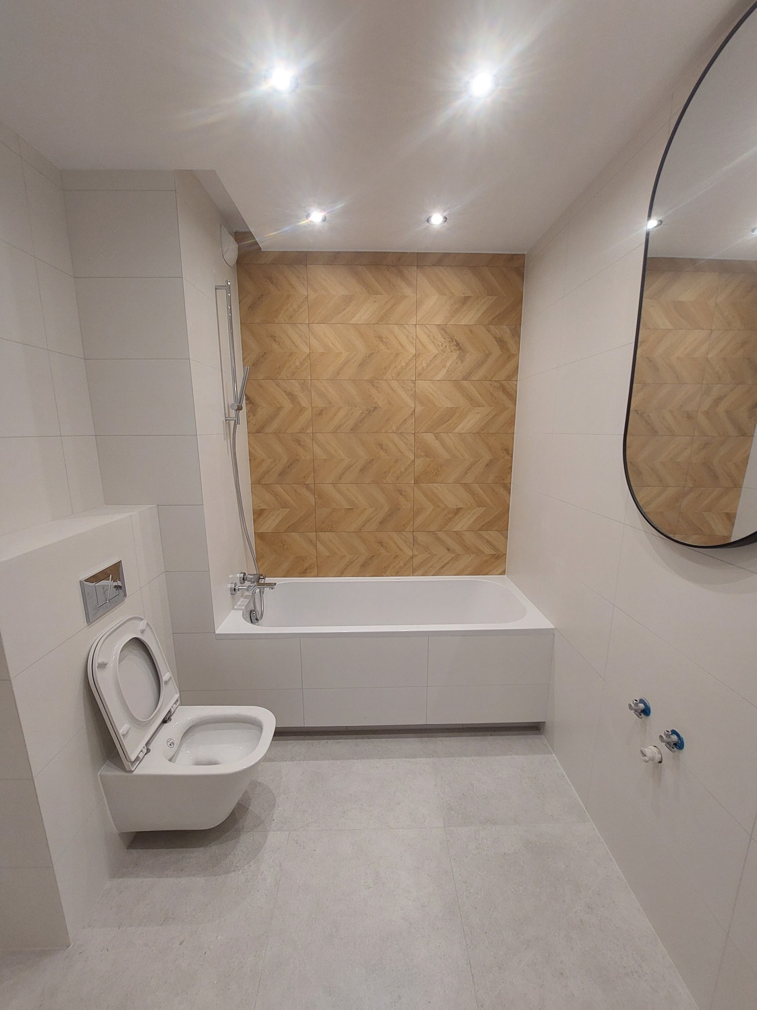 Nowoczesna łazienka z białymi płytkami, akcentem w postaci drewnopodobnej mozaiki nad wanną, wiszącą toaletą i owalnym lustrem.