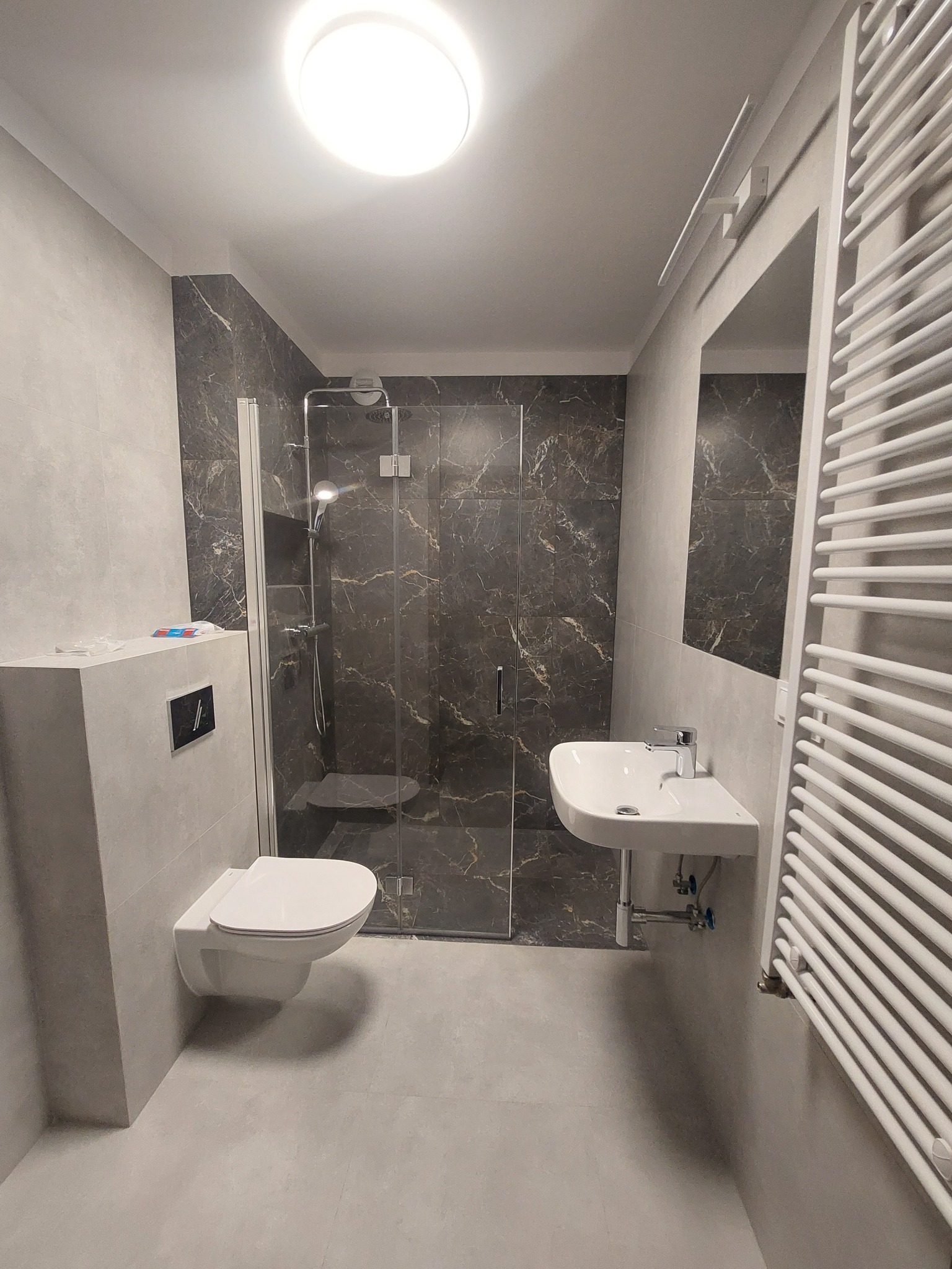 Nowoczesna łazienka z kabiną prysznicową wykończoną płytkami imitującymi marmur, podwieszana toaleta i umywalka, grzejnik drabinkowy.