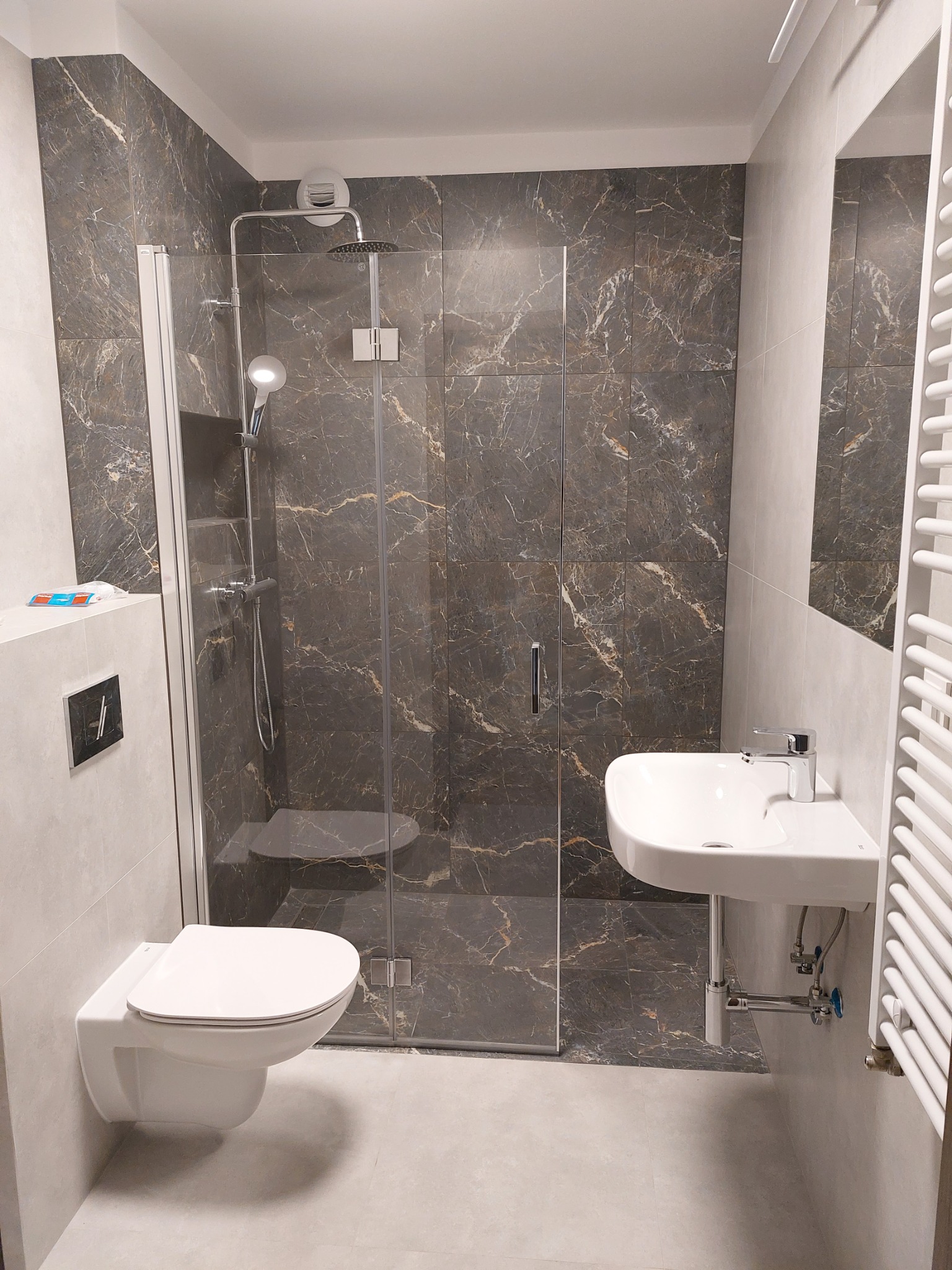 Nowoczesna łazienka z kabiną prysznicową typu walk-in wyłożoną płytkami imitującymi marmur, podwieszaną toaletą i umywalką oraz grzejnikiem drabinkowym.