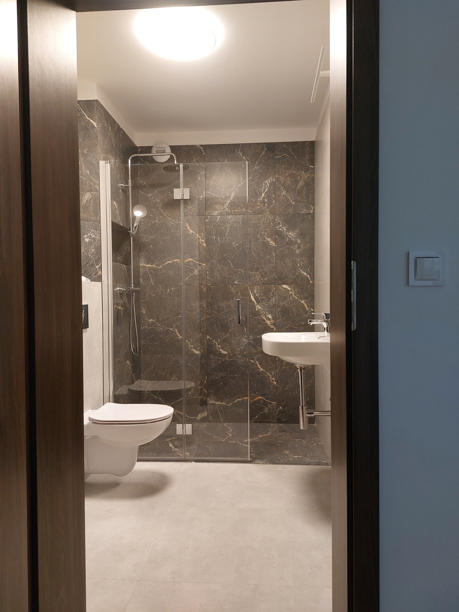 Nowoczesna łazienka z kabiną prysznicową ze szklanymi drzwiami, marmurowymi płytkami na ścianach, wiszącą toaletą i umywalką na postumencie, widziana zza drewnianej futryny drzwi.