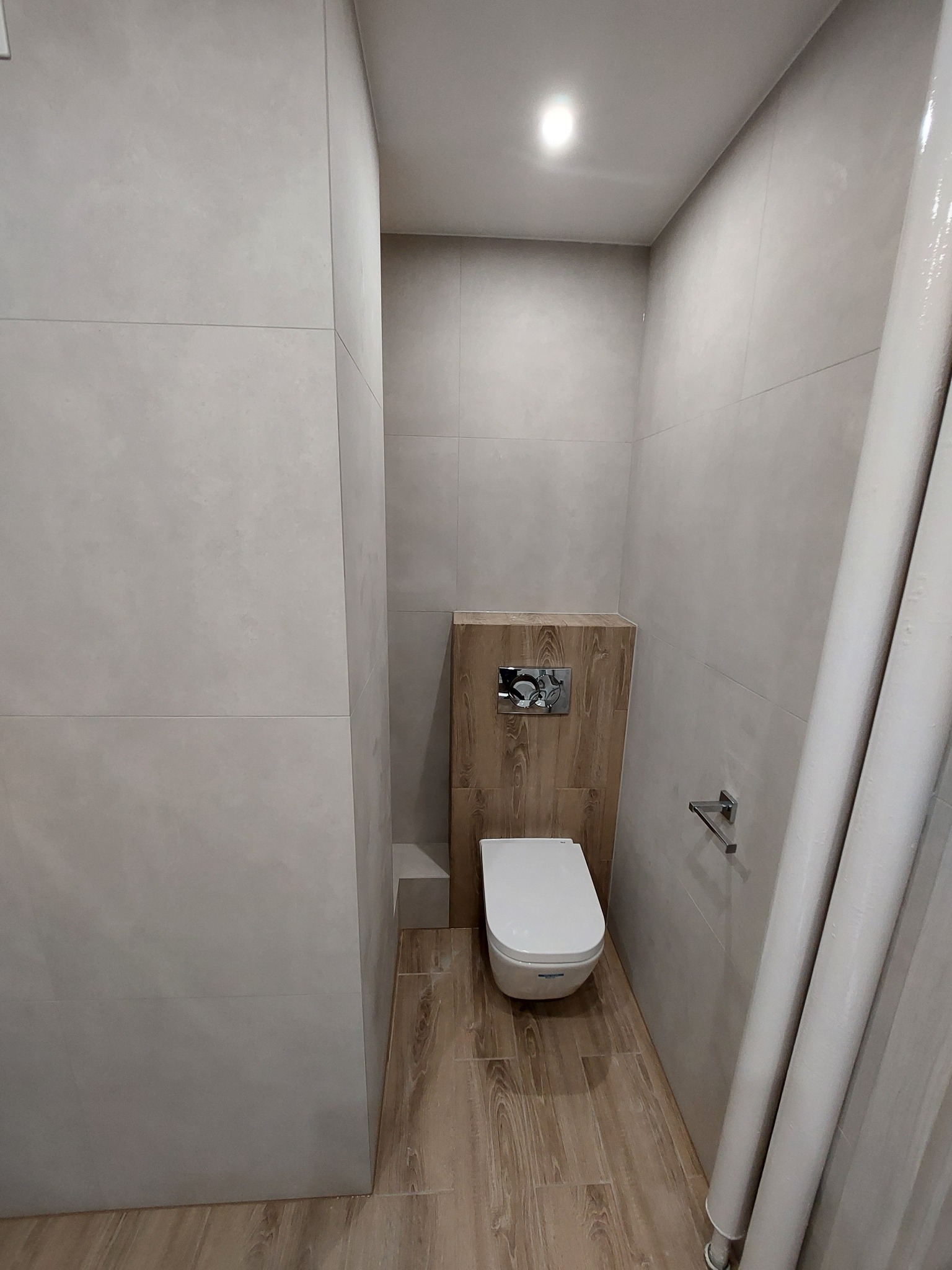 Nowoczesna toaleta podwieszana z drewnianą zabudową stelaża, wykończona płytkami w odcieniach szarości i beżu, z widocznymi rurami kanalizacyjnymi w rogu.