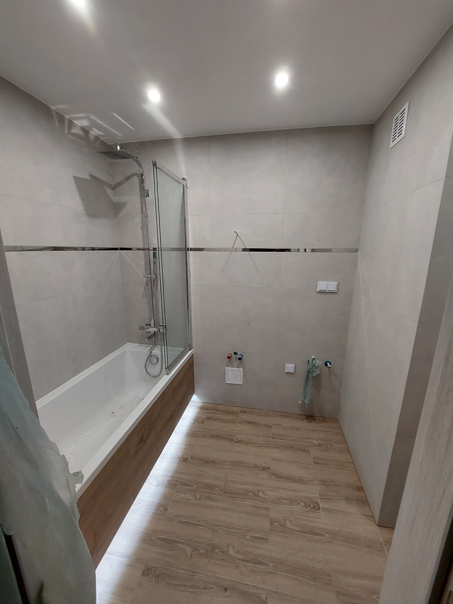 Wykończona łazienka w jasnych odcieniach szarości z drewnopodobną podłogą, widoczna wanna z parawanem nawannowym, chromowana armatura oraz przygotowane przyłącza wodno-kanalizacyjne.