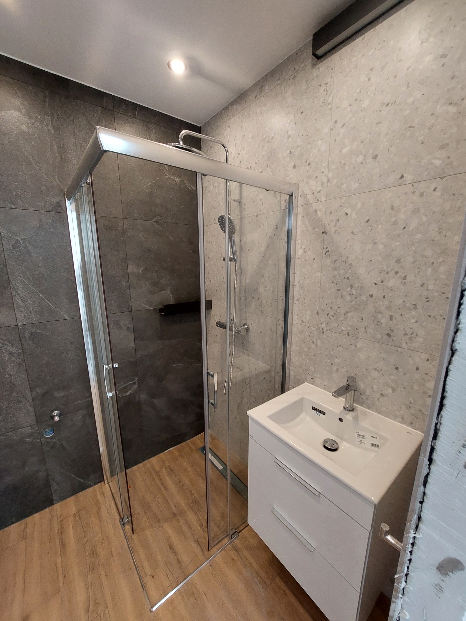 Nowoczesna łazienka po remoncie: kabina prysznicowa z chromowanymi profilami i deszczownicą, umywalka nablatowa z białą szafką, szare płytki imitujące kamień na ścianach i drewnopodobne panele...