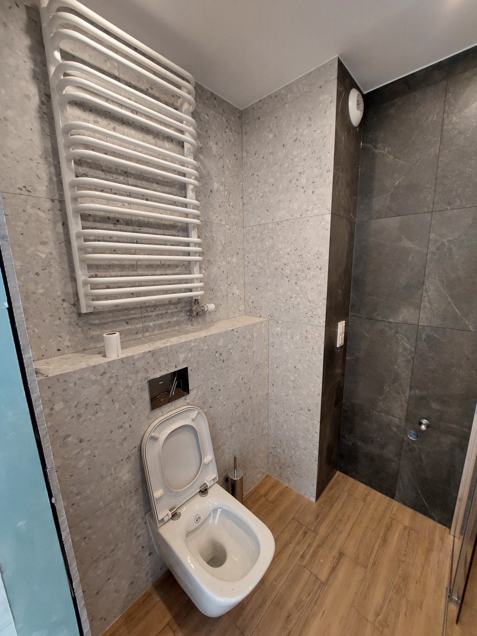 Nowoczesna łazienka z toaletą podwieszaną, grzejnikiem drabinkowym i panelami ściennymi imitującymi lastryko, uzupełniona drewnopodobnymi płytkami podłogowymi i ciemnoszarą strefą prysznicową...