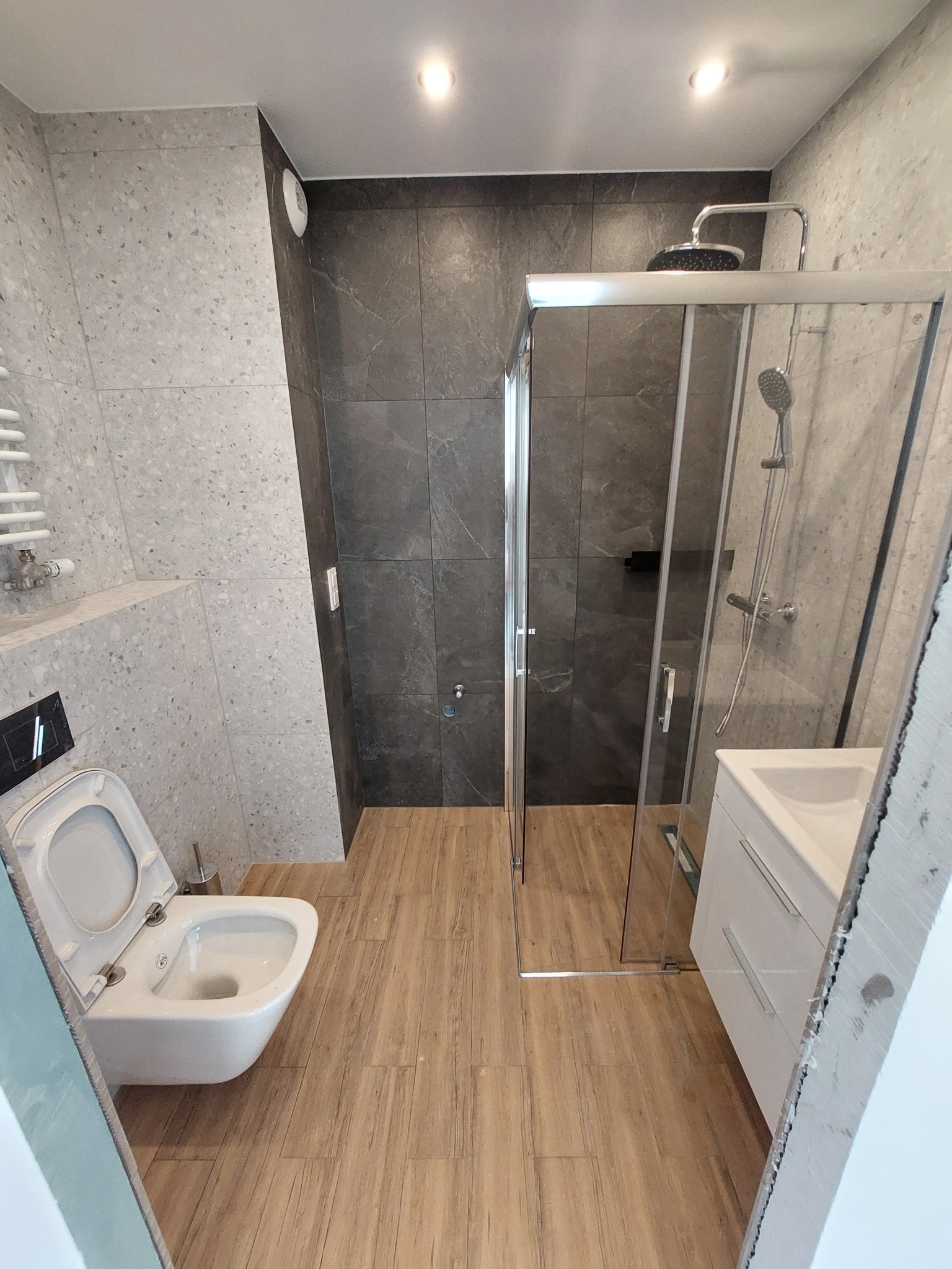 Wykończona łazienka w Wieliczce z kabiną prysznicową, umywalką z szafką, toaletą podwieszaną i grzejnikiem drabinkowym. Ściany wyłożone płytkami w odcieniach szarości, a podłoga drewnopodobnymi...